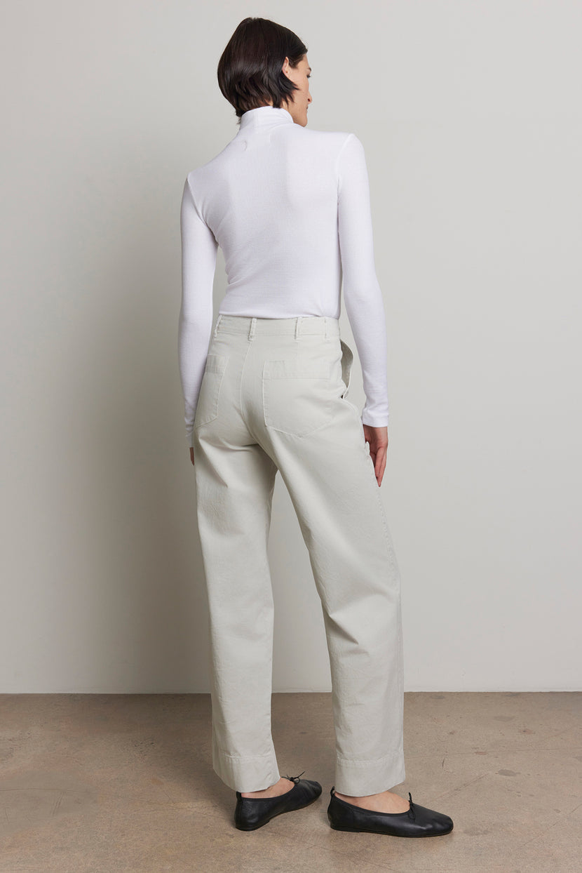 A person stands facing away, donning a white turtleneck and the VENTURA PANT, a pair of light-colored, wide-leg high-waisted pants crafted from cotton twill by Velvet by Jenny Graham. Black shoes complete the utilitarian ensemble against a plain background.