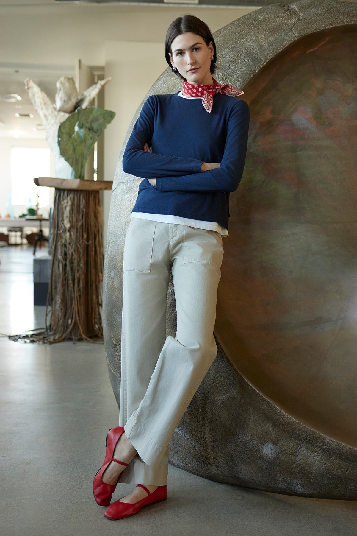   A woman dressed in a navy shirt, Velvet by Jenny Graham's VENTURA PANT featuring wide-leg beige slacks with slash pockets, and accessorized with red shoes and a red scarf stands in a modern room, highlighted by the striking circular sculpture behind her. 