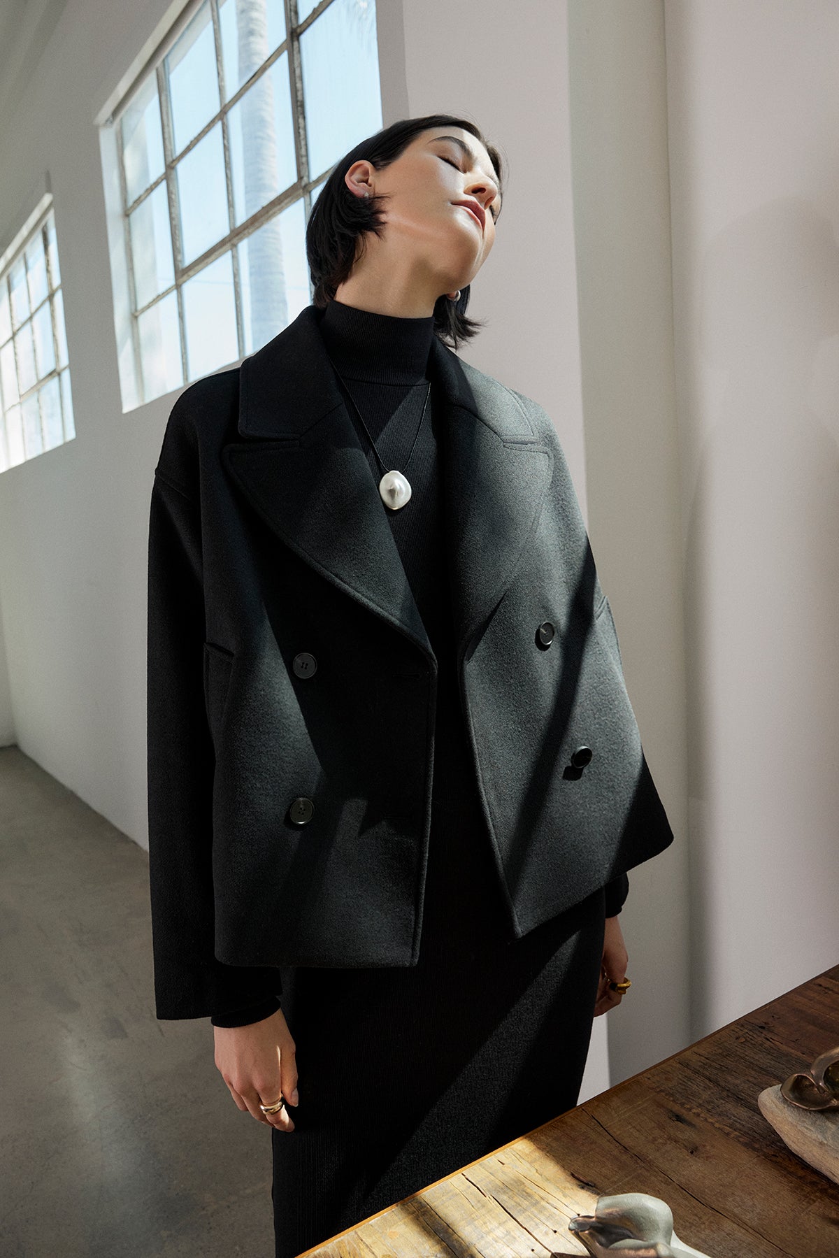   A person in the WESTWOOD PEACOAT by Velvet by Jenny Graham and a dress stands in a bright room, tilting their head back with eyes closed near a wooden table. 