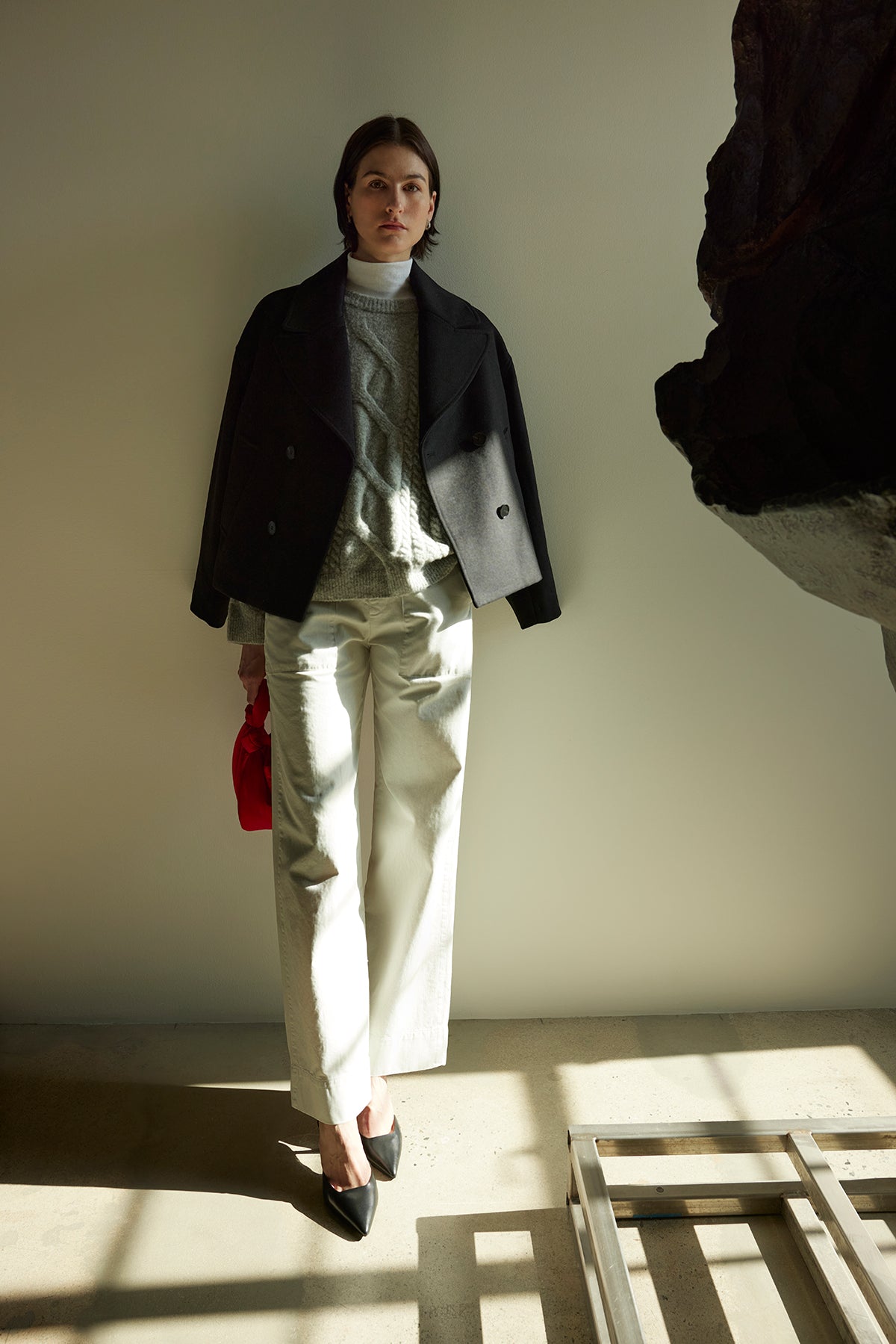   A woman in a Velvet by Jenny Graham WESTWOOD PEACOAT, paired with a gray sweater, white pants, and black shoes, stands against a light wall, holding a small red bag. Her outfit showcases timeless appeal with its classic design and subtle elegance. 