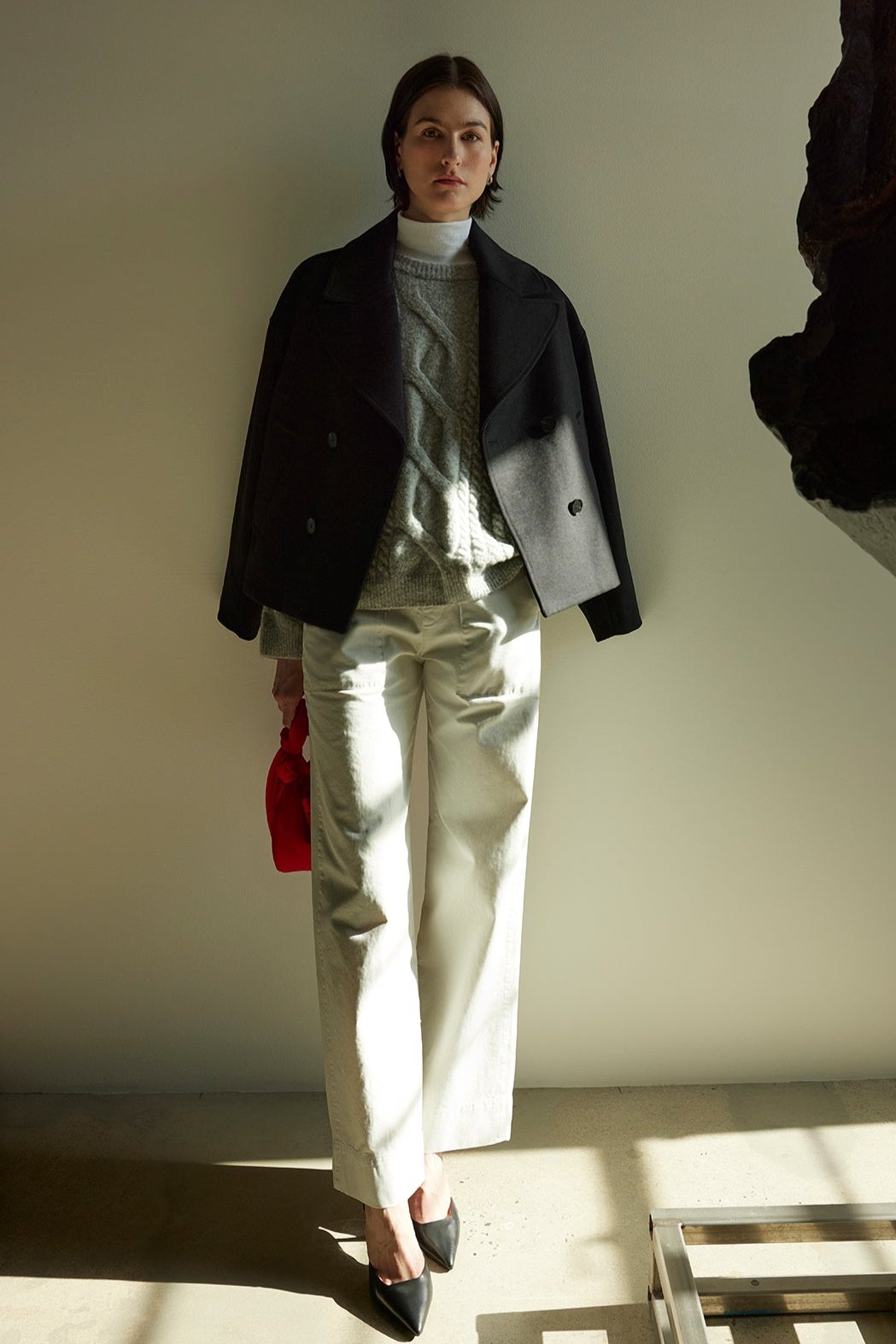   A person stands in a sunlit room wearing a dark jacket, light sweater, and the VENTURA PANT by Velvet by Jenny Graham, made of cotton twill. Black shoes ground the outfit while they hold a red bag next to a large abstract sculpture. 