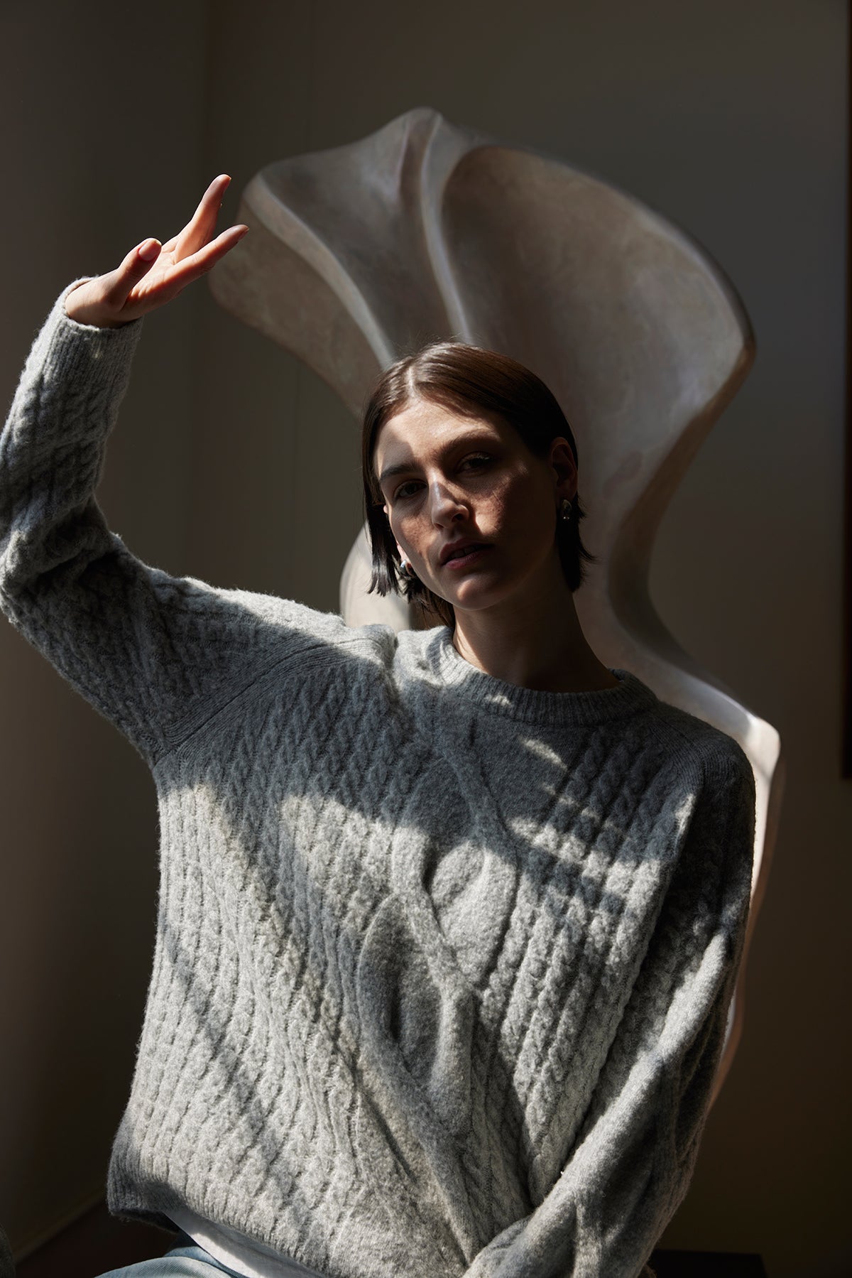   A person wearing the WILSHIRE SWEATER from Velvet by Jenny Graham, a cozy gray wool-yak blend sweater, poses with a peace sign in front of a large abstract sculpture. 