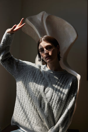 A person wearing the WILSHIRE SWEATER from Velvet by Jenny Graham, a cozy gray wool-yak blend sweater, poses with a peace sign in front of a large abstract sculpture.