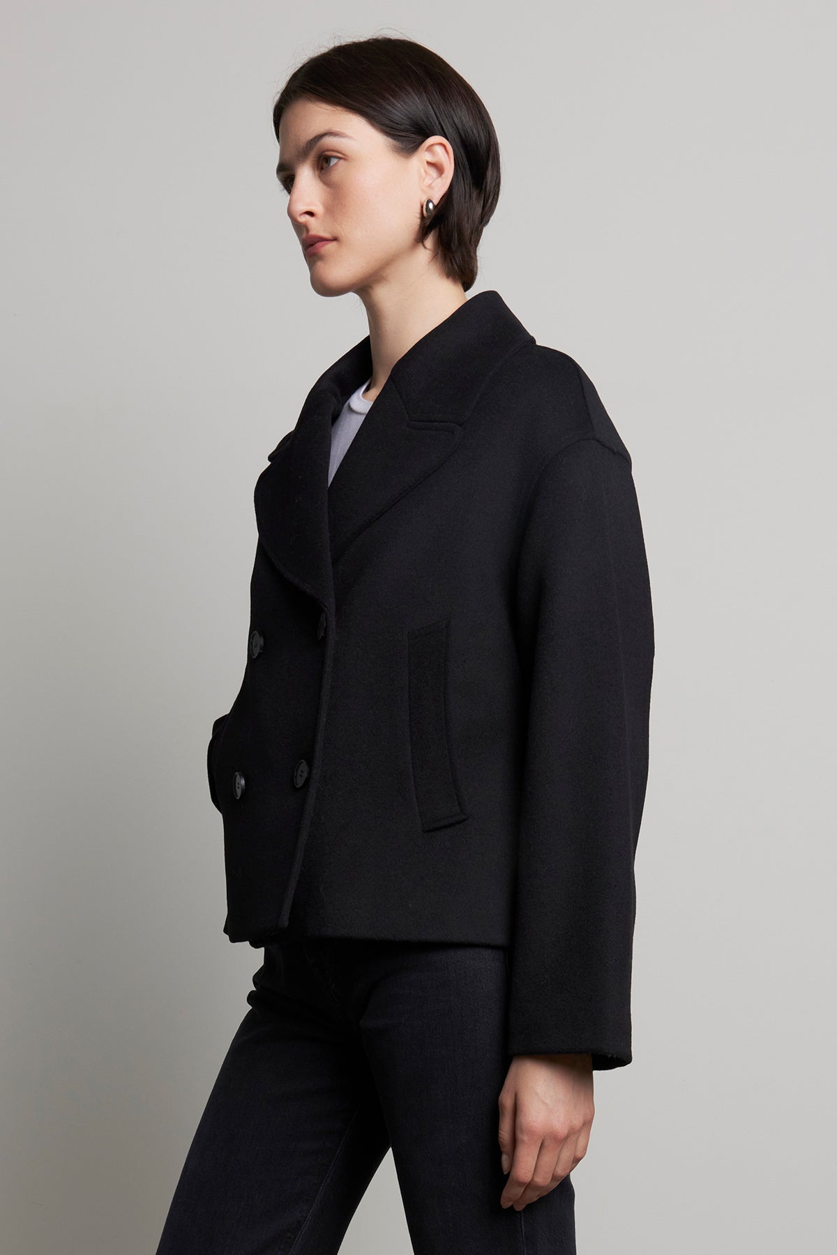   Dressed in the WESTWOOD PEACOAT by Velvet by Jenny Graham, paired with black jeans, a person stands sideways against a plain background, exuding timeless appeal. 