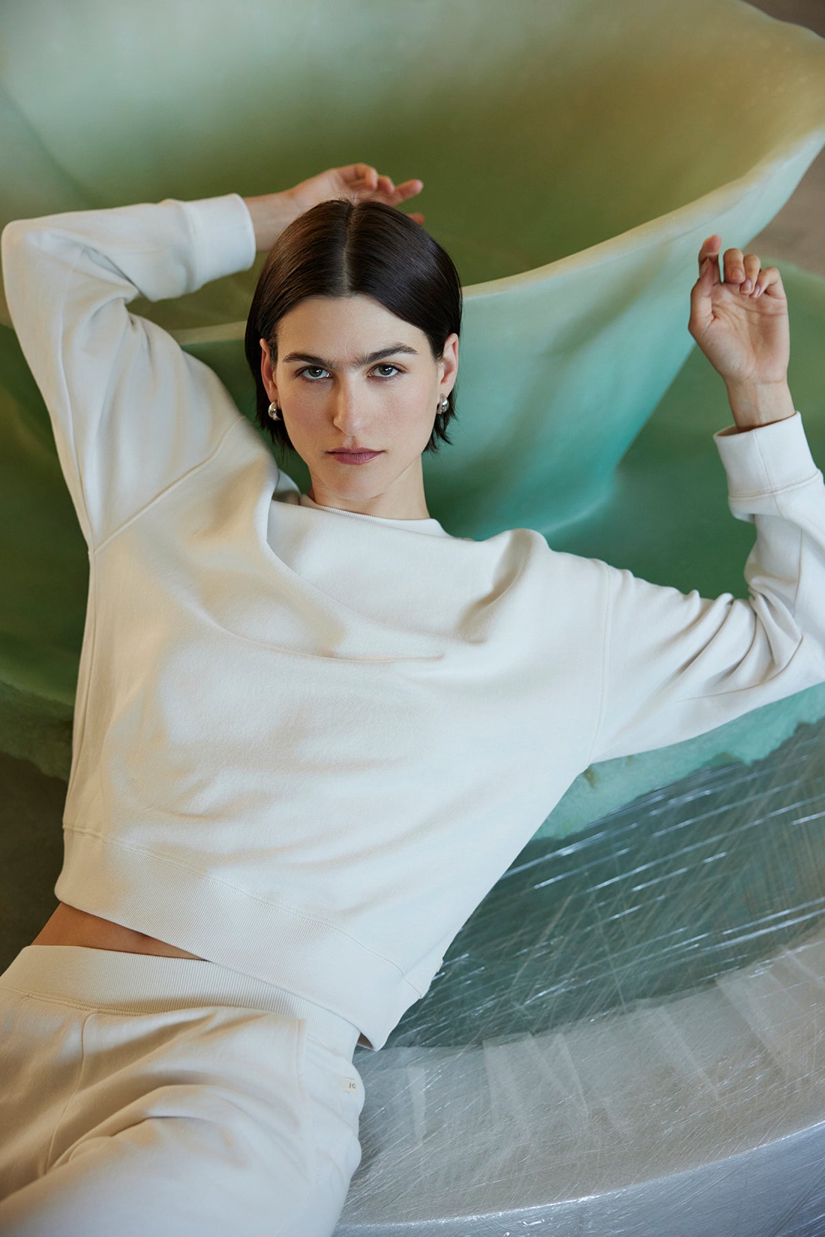   A person wearing the YNEZ SWEATSHIRT by Velvet by Jenny Graham reclines against a large green abstract shape, gazing directly at the camera. 