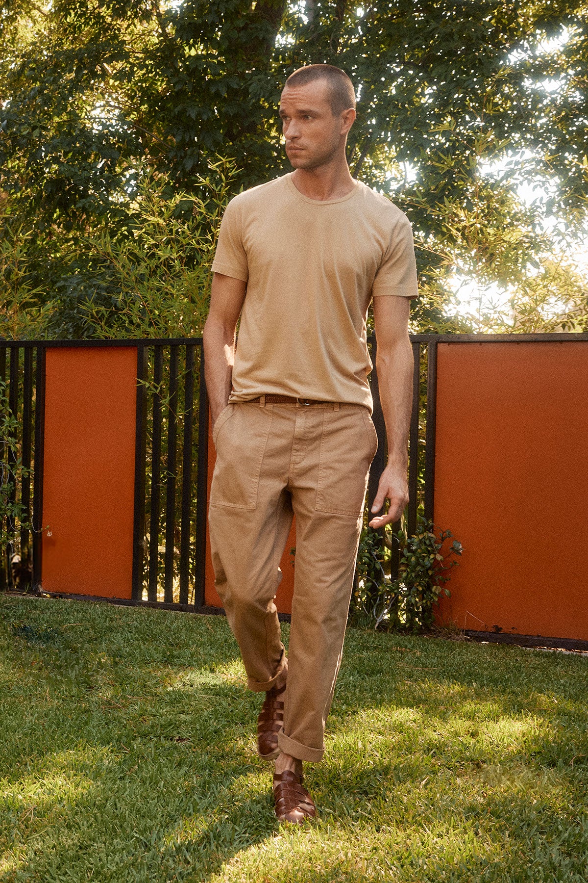   A man in a vintage feel Howard Tee by Velvet by Graham & Spencer and beige pants walks on grass in a garden, with a black and red fence and green foliage in the background. 