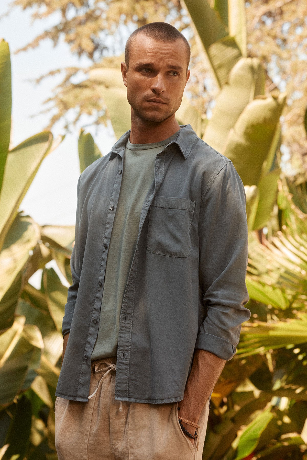   A man wearing a WOLF BUTTON-UP SHIRT by Velvet by Graham & Spencer, specifically a blue button-up over a green t-shirt, stands among large green plants, looking off to the side. 