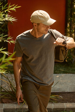 A man wearing a beige cap, Velvet by Graham & Spencer's STITCH TEE in gray, and olive pants carries a backpack over his shoulder while walking outdoors.