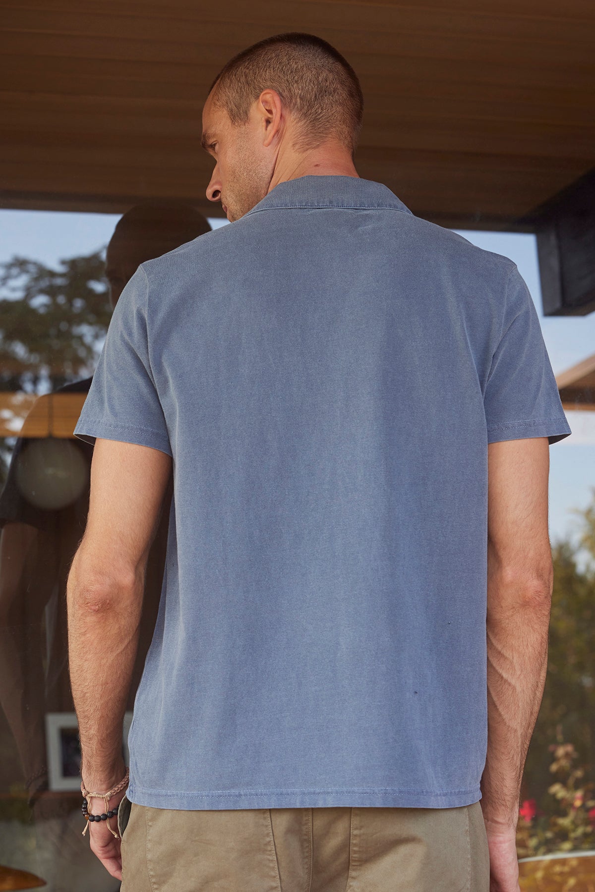   A man with short hair, wearing the stylish ROWAN POLO in a blue, pigment-dyed fabric by Velvet by Graham & Spencer and beige pants, is standing with his back to the camera. 