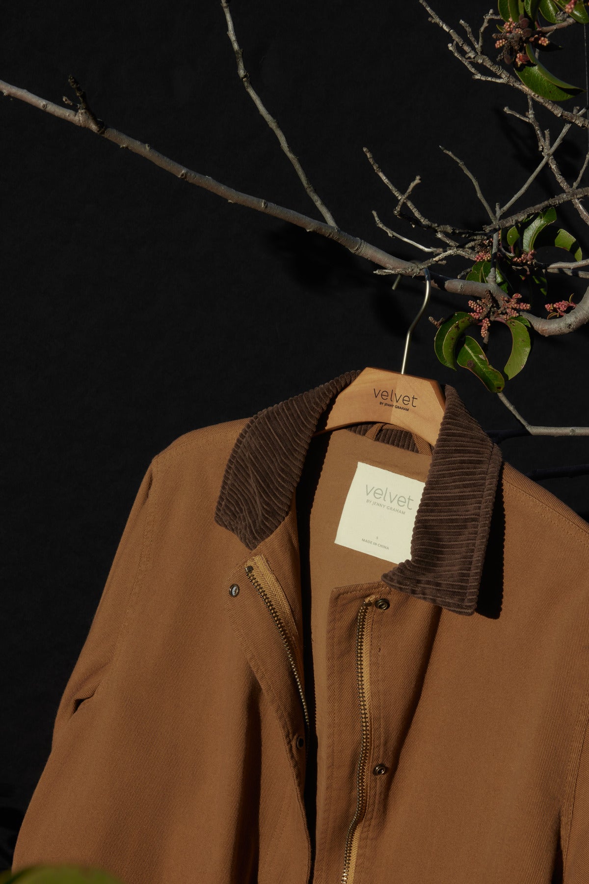   The OAKLEY JACKET, crafted from cotton twill fabric with corduroy trims and a corduroy collar, hangs on a Velvet by Jenny Graham branded hanger. The setting features a dark background adorned with branches and green leaves. 