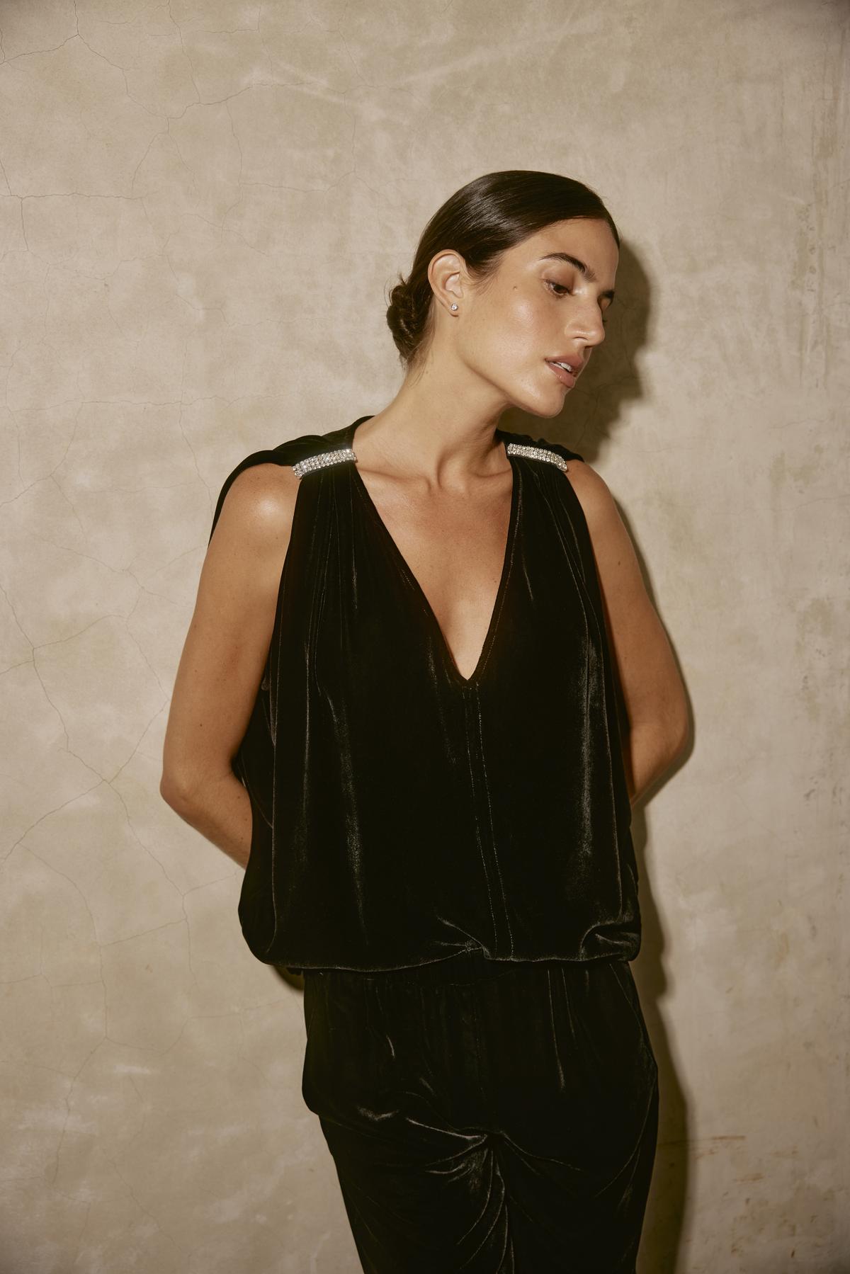   A person with dark hair is standing against a textured wall, wearing the ADELLE SILK VELVET TOP by Velvet by Graham & Spencer. This black silk velvet sleeveless top features decorative straps and a chic v-neckline, making it perfect for holiday gatherings. 