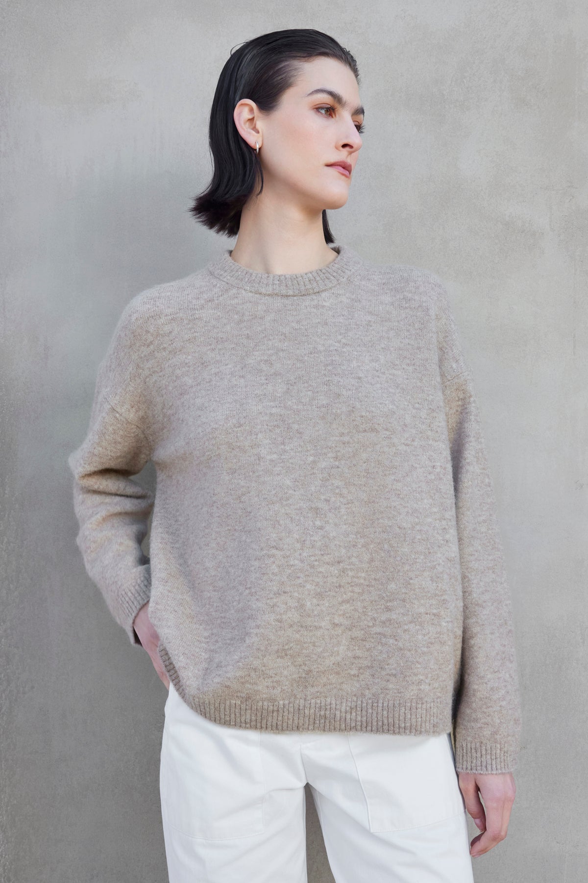   A person with short dark hair is standing against a gray wall, wearing the LAKEWOOD ALPACA SWEATER by Velvet by Jenny Graham and white pants, looking off to the side. 