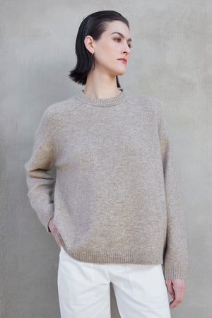 A person with short dark hair is standing against a gray wall, wearing the LAKEWOOD ALPACA SWEATER by Velvet by Jenny Graham and white pants, looking off to the side.