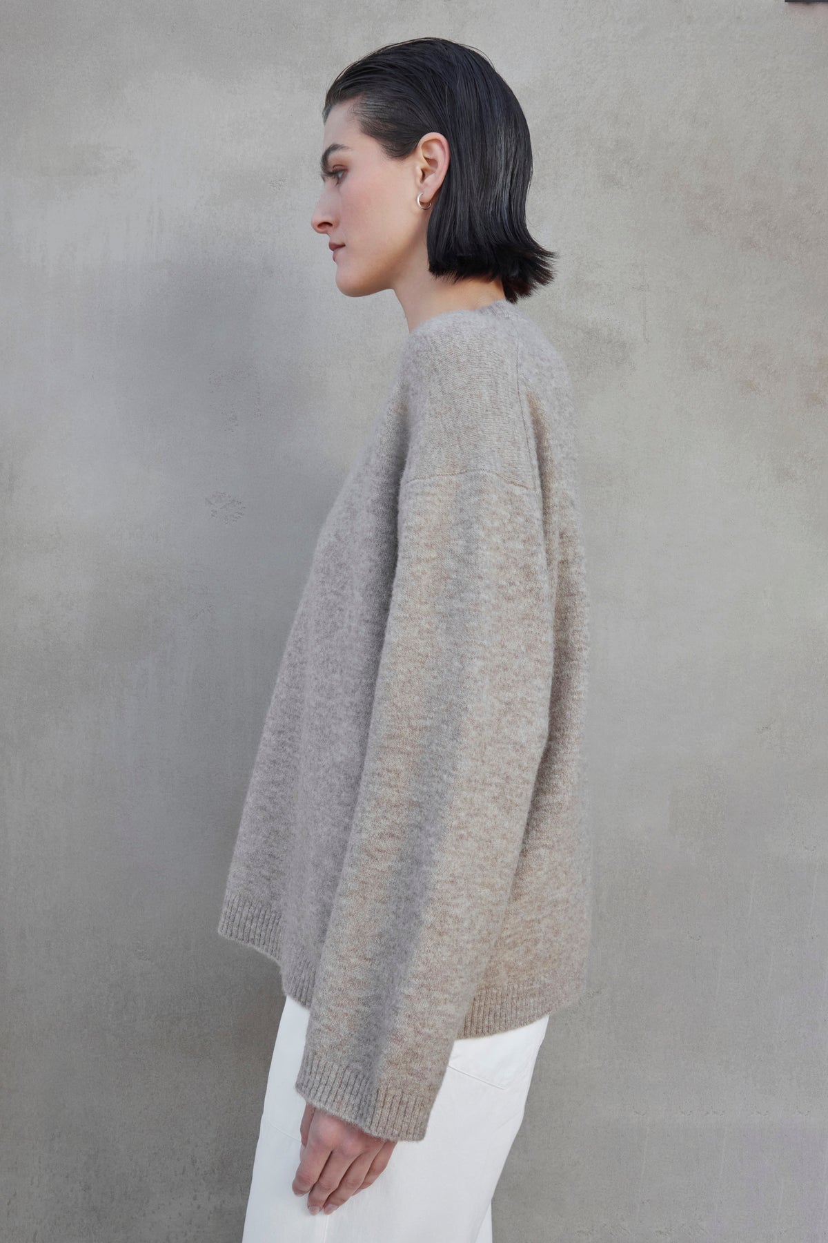   A person with short, dark hair is standing in profile against a plain, neutral-colored background, wearing the LAKEWOOD ALPACA SWEATER by Velvet by Jenny Graham and white pants. 