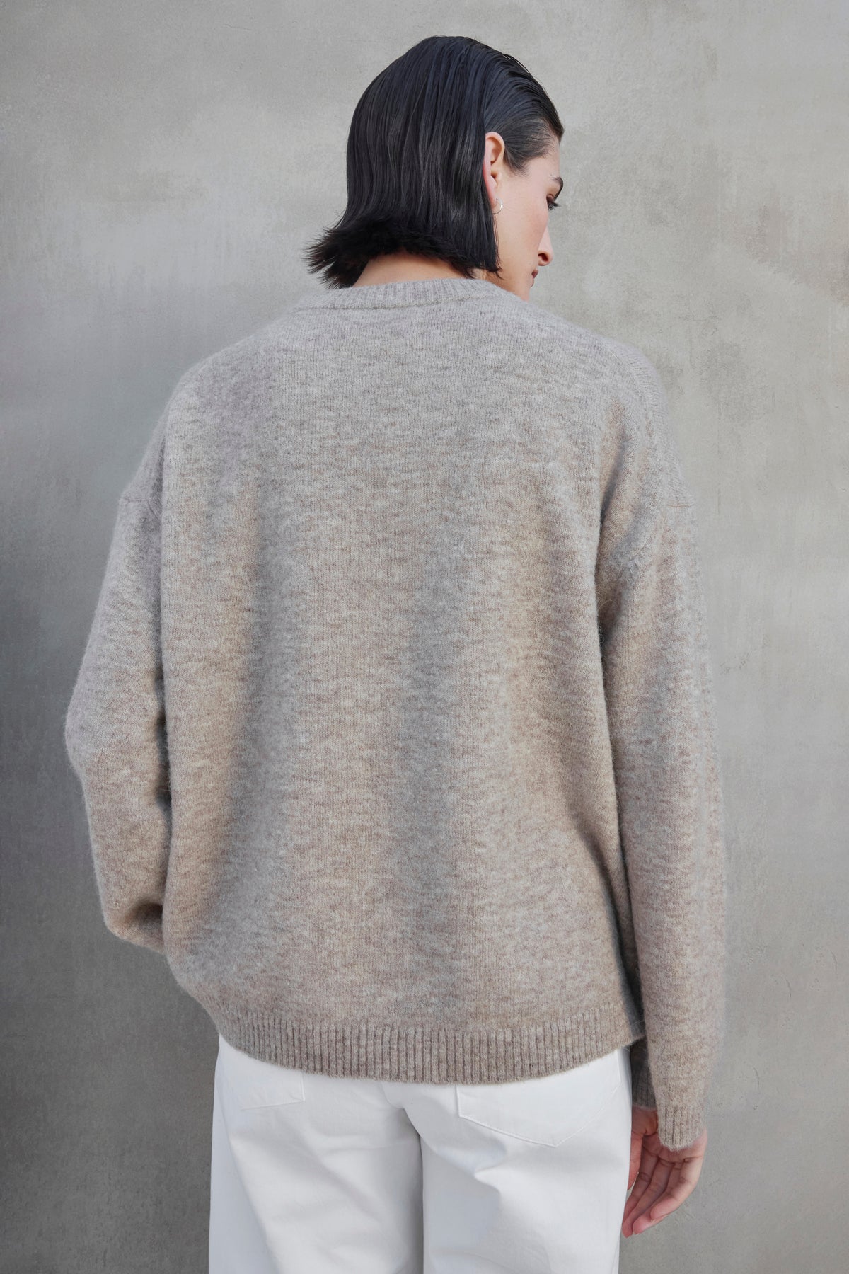   A person with short dark hair, wearing the LAKEWOOD ALPACA SWEATER by Velvet by Jenny Graham and white pants, is standing with their back to the camera against a gray background. 