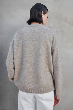 A person with short dark hair, wearing the LAKEWOOD ALPACA SWEATER by Velvet by Jenny Graham and white pants, is standing with their back to the camera against a gray background.