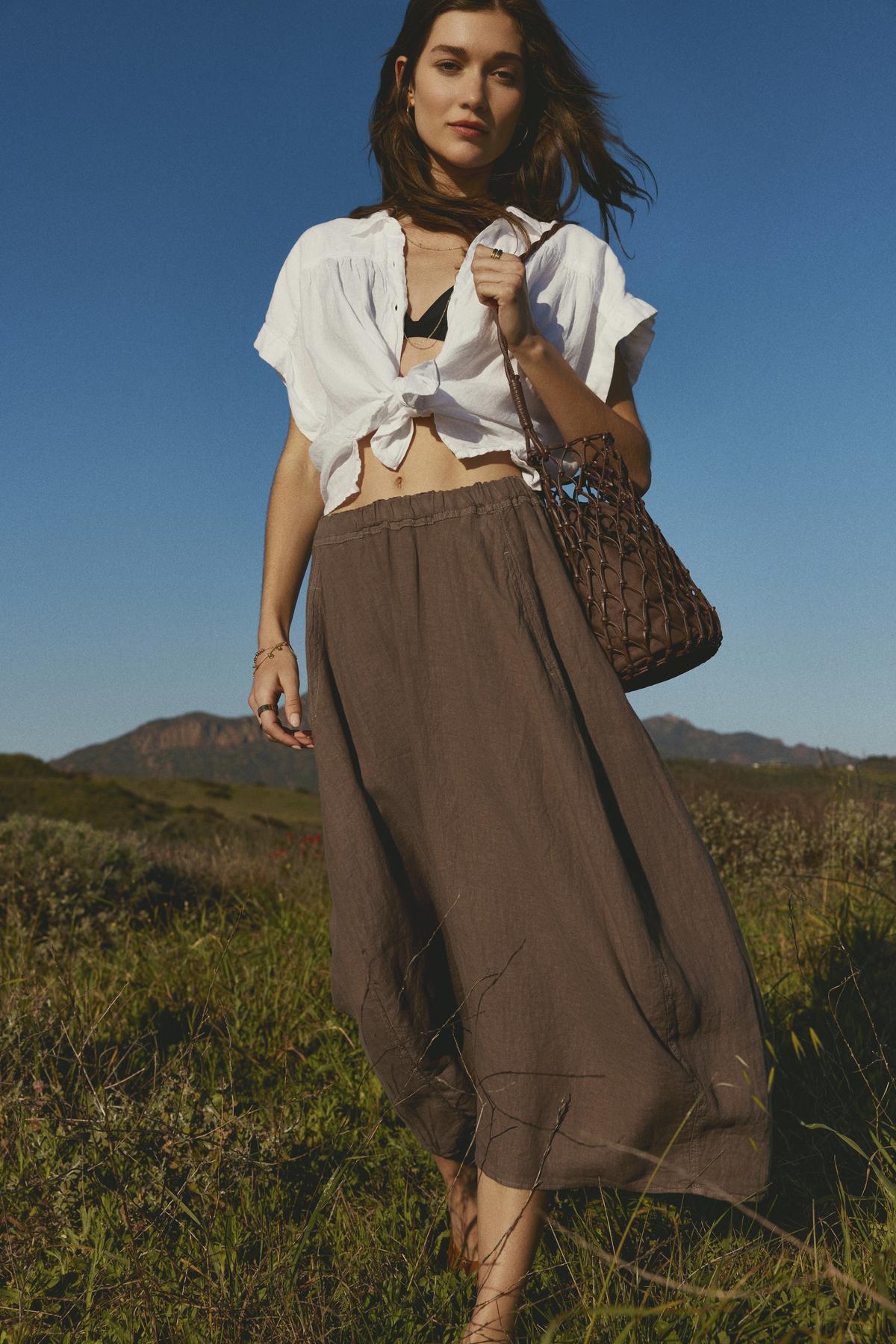 A woman stands in a field wearing the ARIA LINEN TOP by Velvet by Graham & Spencer, paired with a brown skirt and holding a woven bag under the clear blue sky.-36443419738305