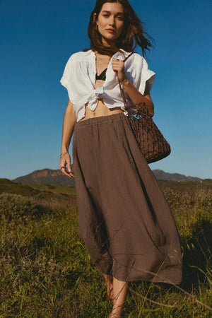 A woman stands in a field wearing the ARIA LINEN TOP by Velvet by Graham & Spencer, paired with a brown skirt and holding a woven bag under the clear blue sky.