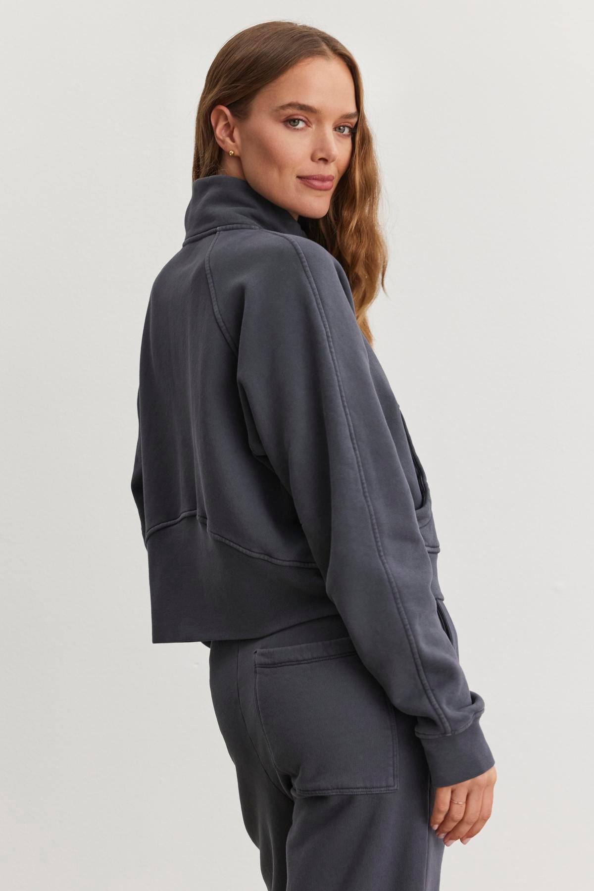   A woman wearing the Velvet by Graham & Spencer Cecilia Full-Zip Jacket in dark gray stands sideways, her long brown hair cascading down, against a plain background. 