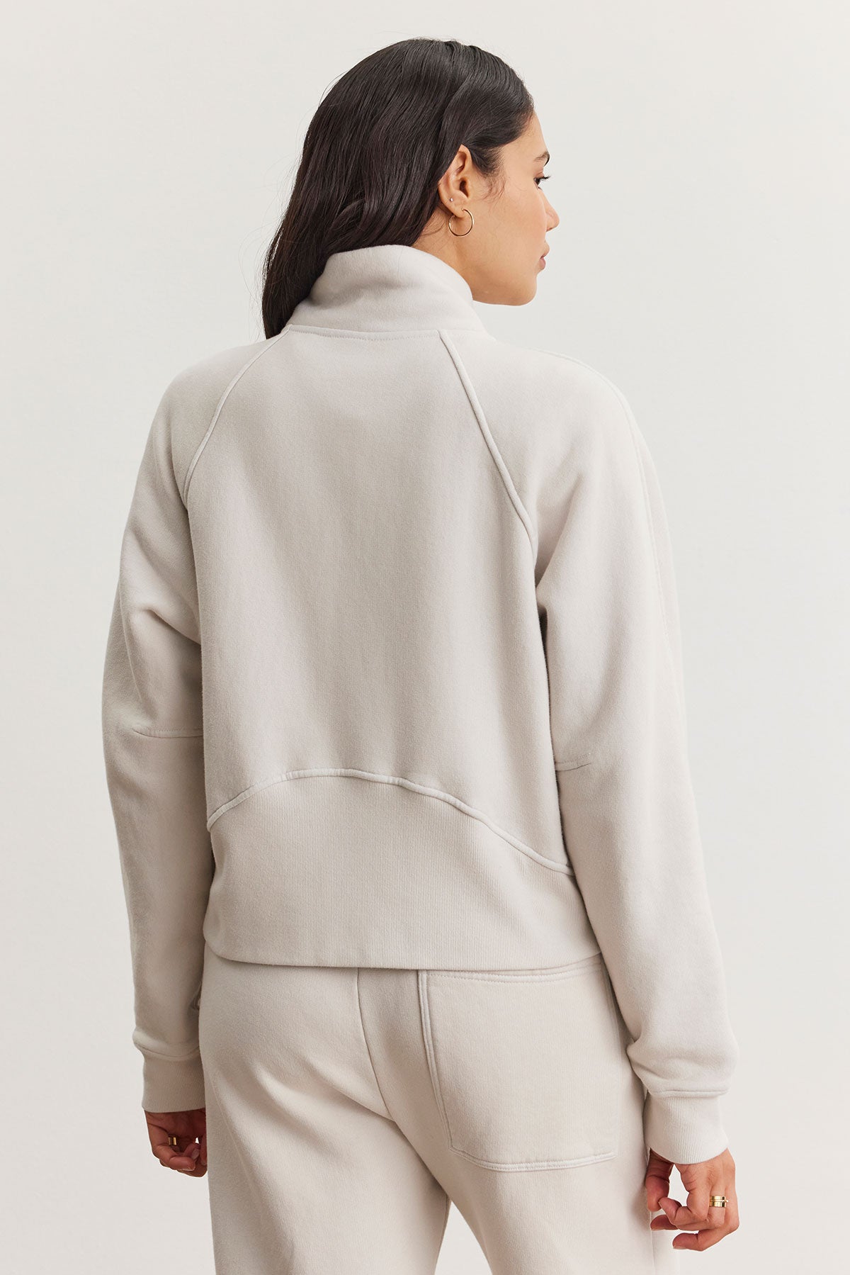   A woman is wearing the Velvet by Graham & Spencer CECILIA FULL-ZIP JACKET, a cozy essential in light gray cotton fleece with raglan sleeves, accompanied by matching pants, as she faces away on a plain background. 