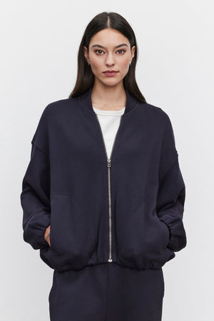 A woman with long dark hair wears the GENEVIEVE ZIP-UP SWEATSHIRT from Velvet by Graham & Spencer over a white top. She stands against a plain white background, looking directly at the camera with her hands in her sweatshirt pockets. The navy blue sweatshirt has an oversized fit, giving it a relaxed and comfy look.