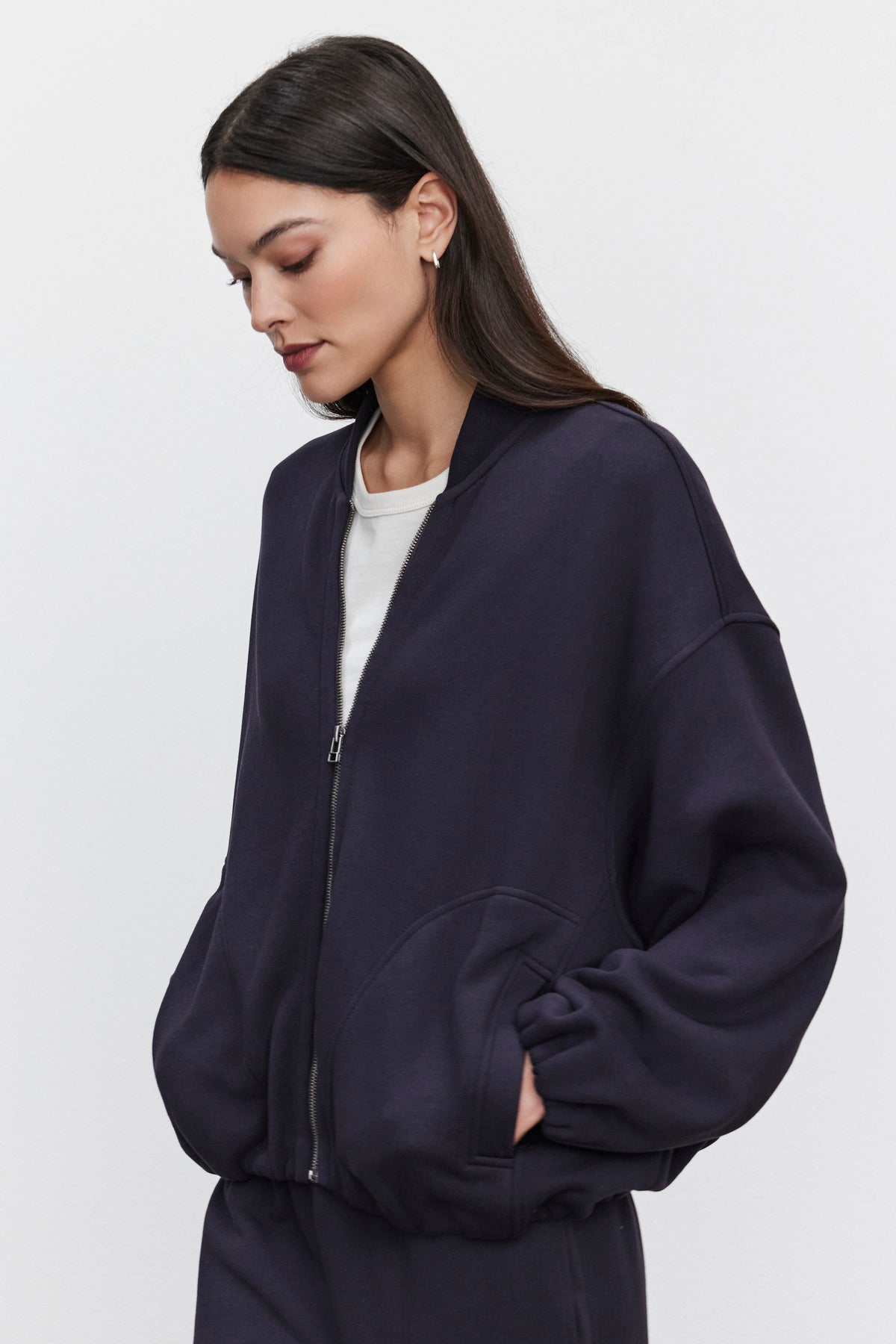   A woman with long dark hair is wearing the GENEVIEVE ZIP-UP SWEATSHIRT by Velvet by Graham & Spencer in navy blue, along with an oversized fit over a white top. She stands against a plain background with her hands in her jacket pockets. 