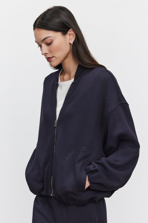 A woman with long dark hair is wearing the GENEVIEVE ZIP-UP SWEATSHIRT by Velvet by Graham & Spencer in navy blue, along with an oversized fit over a white top. She stands against a plain background with her hands in her jacket pockets.