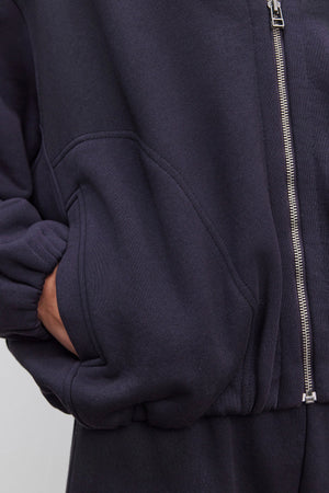 A person wearing an oversized fit, dark GENEVIEVE ZIP-UP SWEATSHIRT by Velvet by Graham & Spencer with their hand in the pocket. The sweatshirt, made from heavier-weight cotton fleece, features a visible zipper and elasticized cuffs.