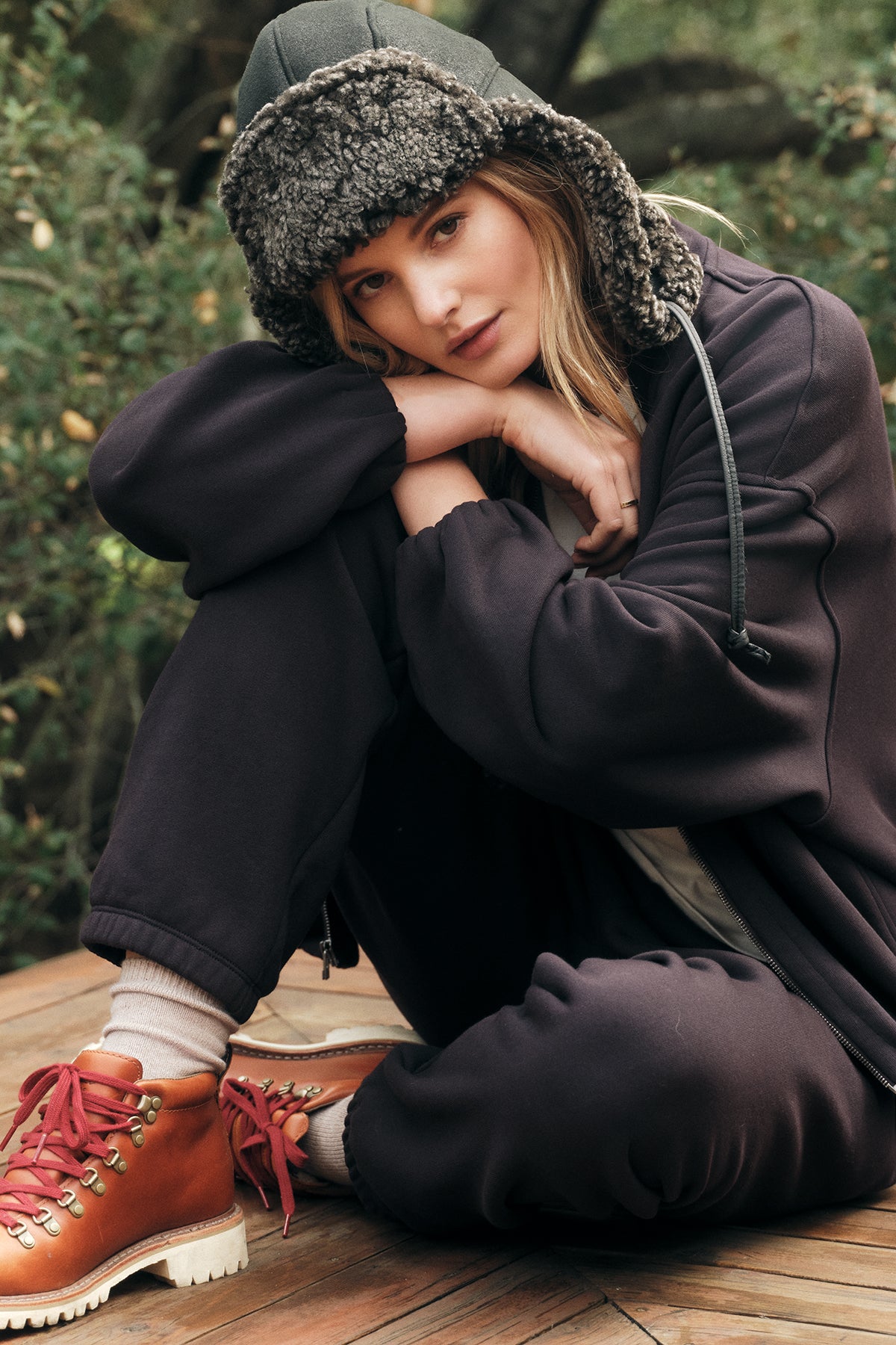   A woman sits outside on a wooden surface, wearing dark sweatpants and the Velvet by Graham & Spencer GENEVIEVE ZIP-UP SWEATSHIRT in an oversized fit, complemented by a wool-lined hat and brown boots with red laces, resting her head on her hand. Green plants are visible behind her. 