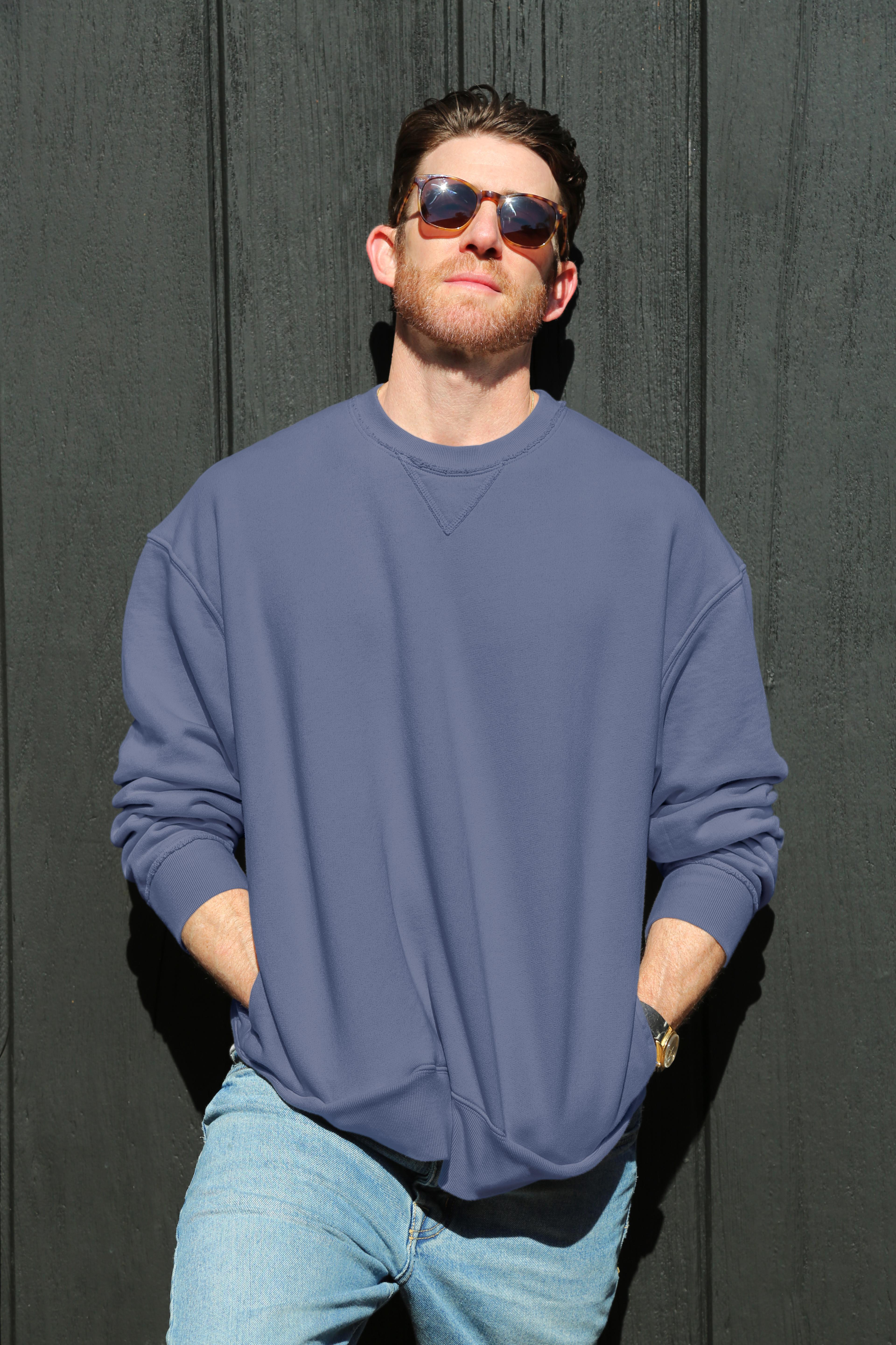   A man in sunglasses stands with hands in his pockets, wearing the Velvet by Graham & Spencer FORREST SWEATSHIRT, a relaxed fit cotton fleece, paired with jeans and leaning against a dark wall. 