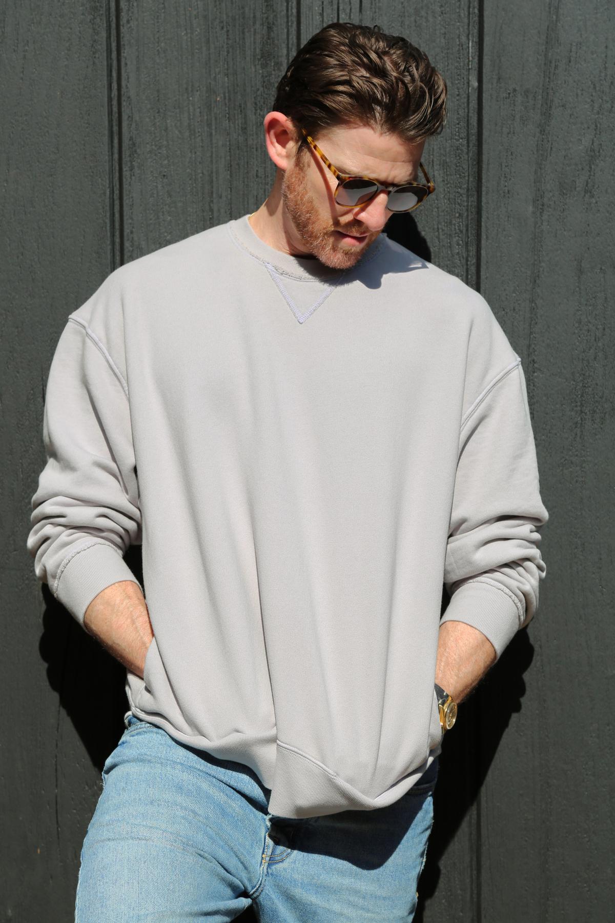   A man in a FORREST SWEATSHIRT by Velvet by Graham & Spencer, made from garment-dyed cotton fleece, paired with blue jeans stands against a dark wall. He has his hands in his pockets, is wearing sunglasses, and is looking down. 