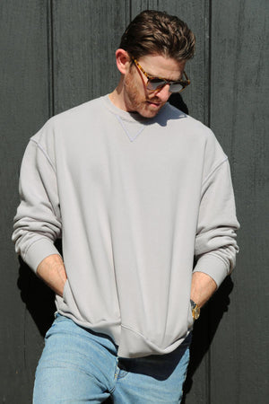 A man in a FORREST SWEATSHIRT by Velvet by Graham & Spencer, made from garment-dyed cotton fleece, paired with blue jeans stands against a dark wall. He has his hands in his pockets, is wearing sunglasses, and is looking down.