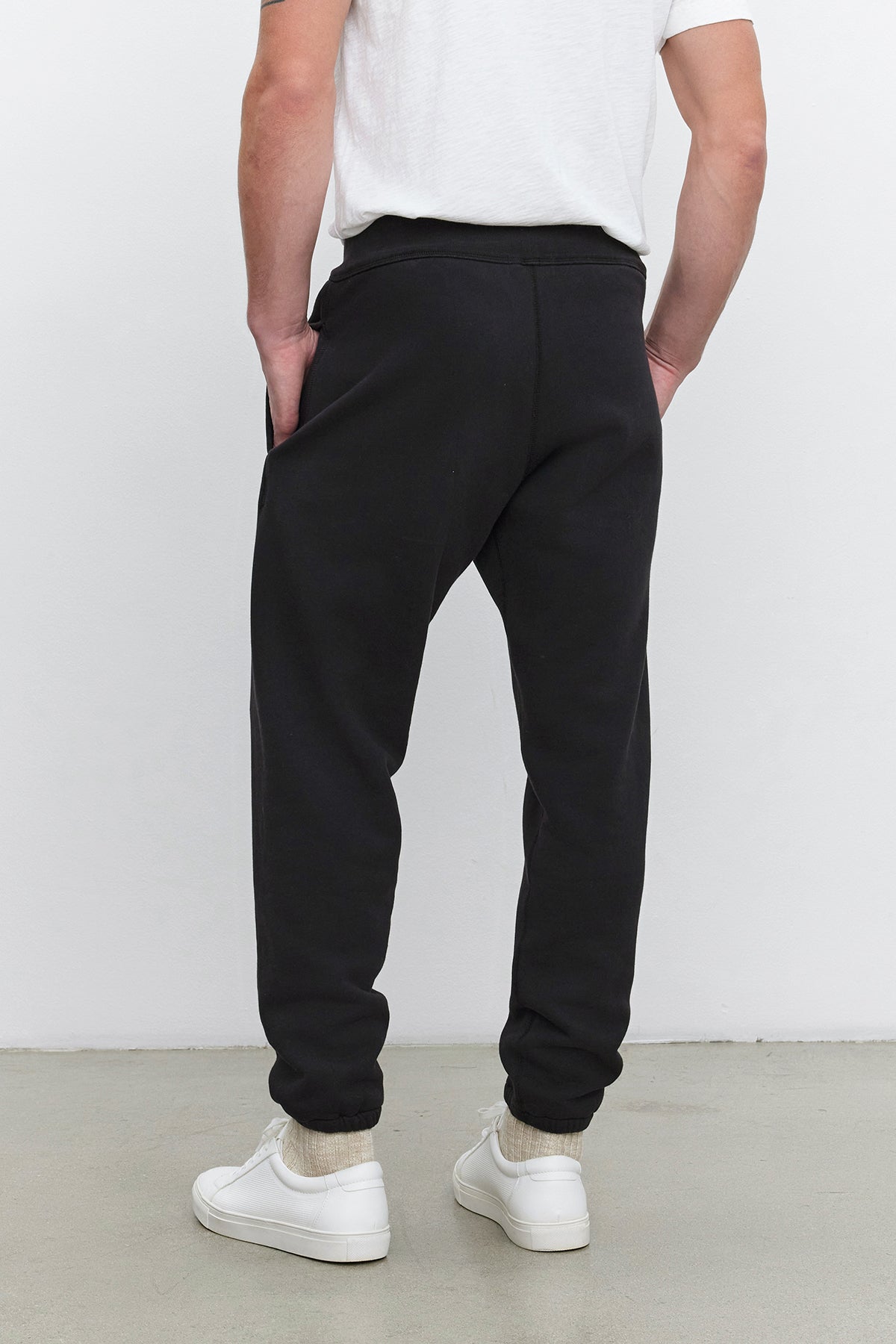   Rear view of a person wearing a KENJI SWEATPANT by Velvet by Graham & Spencer, styled with a white T-shirt, white socks, and white sneakers, standing against a white background. The comfortable fit is highlighted with hands in the pockets of the joggers. 