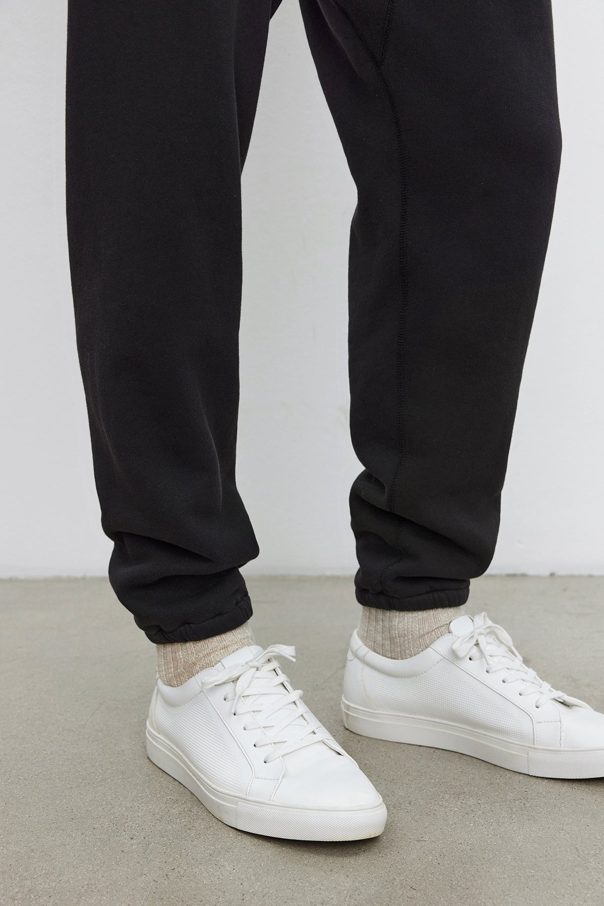   A person wearing KENJI SWEATPANT by Velvet by Graham & Spencer, beige socks, and white sneakers standing on a gray concrete floor, showcasing a comfortable fit that beautifully complements their vintage look. 