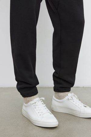 A person wearing KENJI SWEATPANT by Velvet by Graham & Spencer, beige socks, and white sneakers standing on a gray concrete floor, showcasing a comfortable fit that beautifully complements their vintage look.