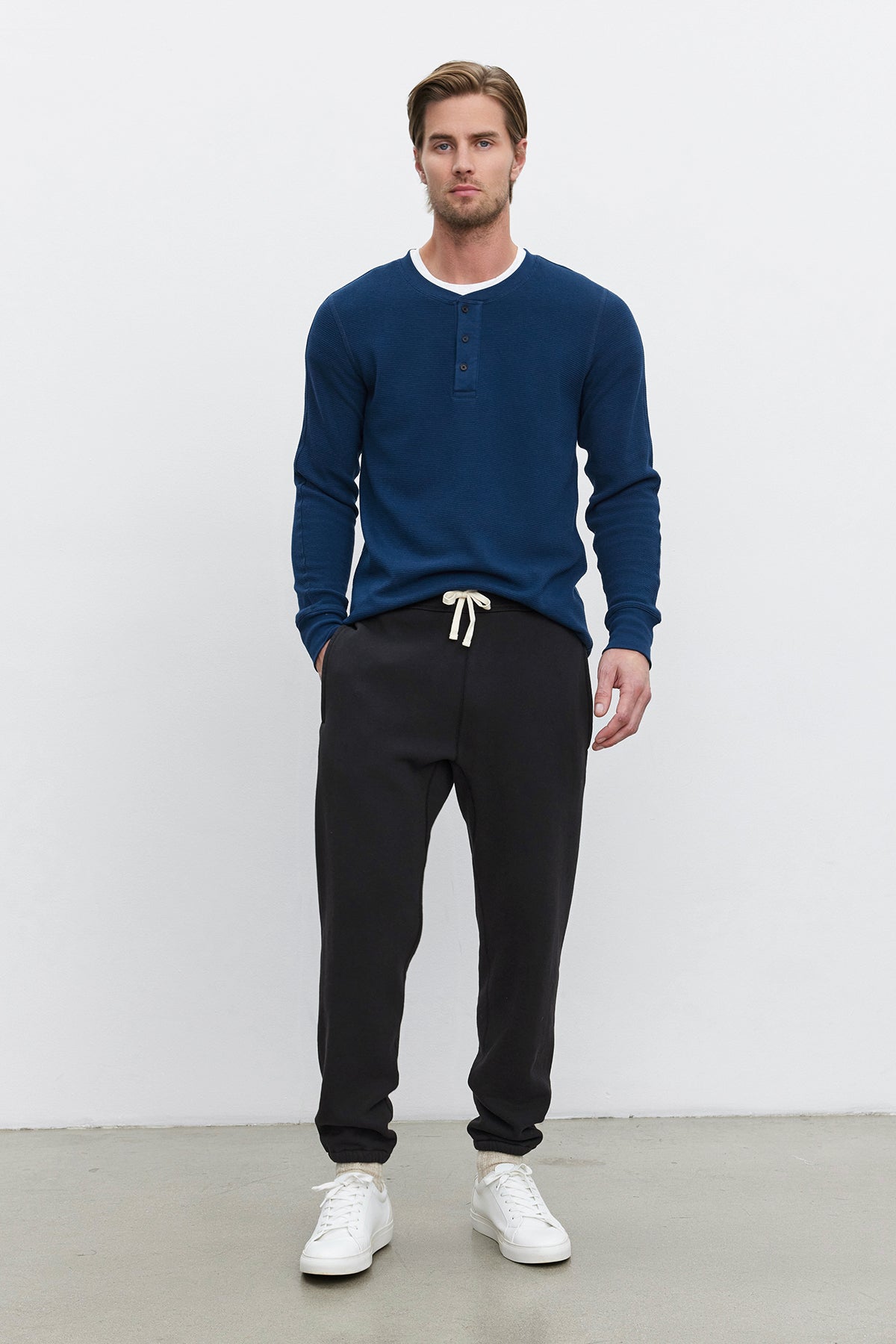 A man stands against a plain white background wearing a blue long-sleeve henley shirt, KENJI SWEATPANT from Velvet by Graham & Spencer in black with a white drawstring, and white sneakers. The sweatpants offer a comfortable fit with their heavier-weight cotton fleece, giving the ensemble a cozy, vintage look.-37893763662017