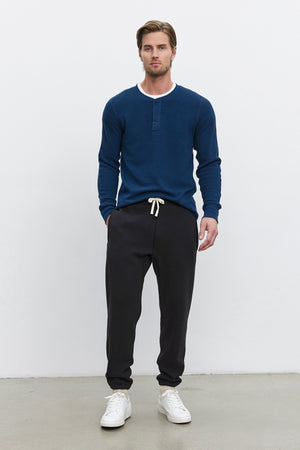 A man stands against a plain white background wearing a blue long-sleeve henley shirt, KENJI SWEATPANT from Velvet by Graham & Spencer in black with a white drawstring, and white sneakers. The sweatpants offer a comfortable fit with their heavier-weight cotton fleece, giving the ensemble a cozy, vintage look.