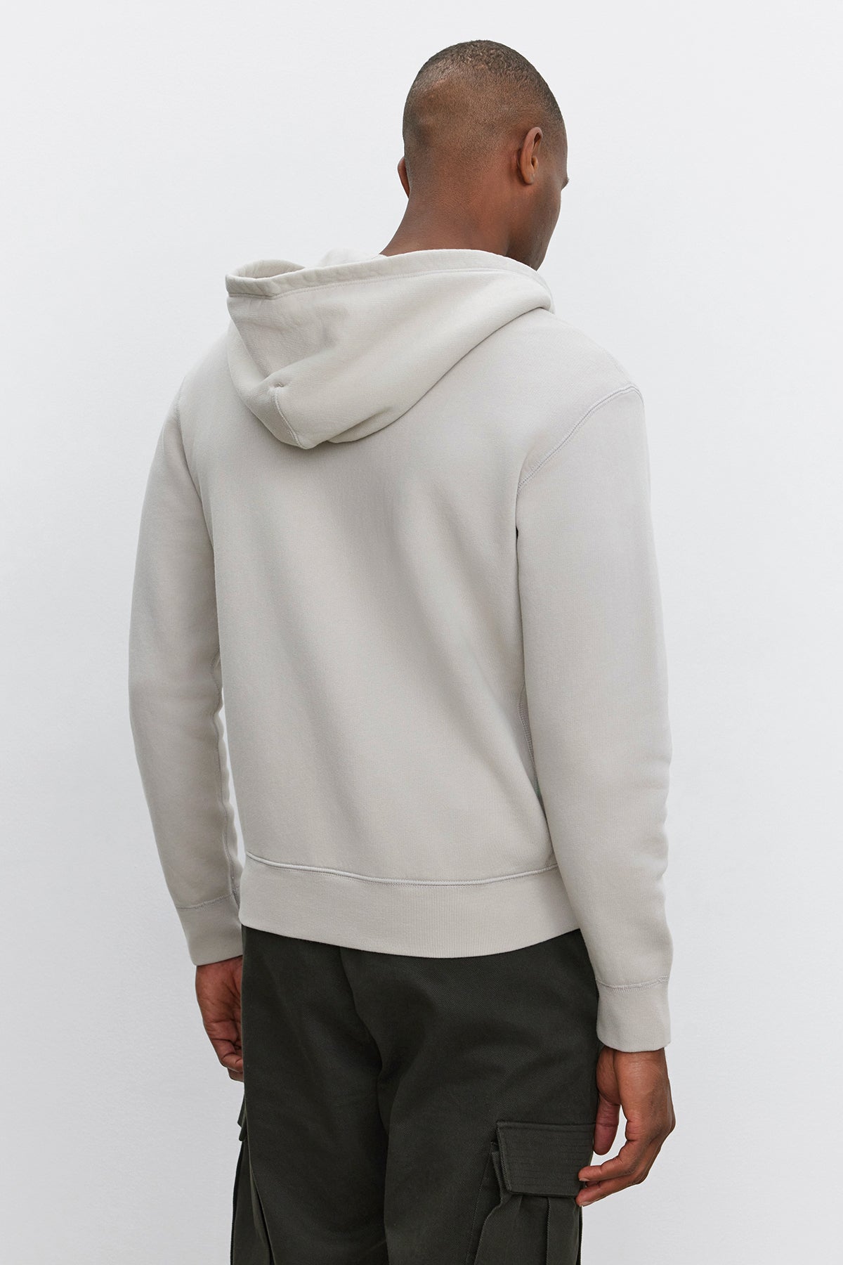   A person stands facing away, wearing a light gray REN HOODIE by Velvet by Graham & Spencer and dark cargo pants against a plain background, giving off a vintage look. 