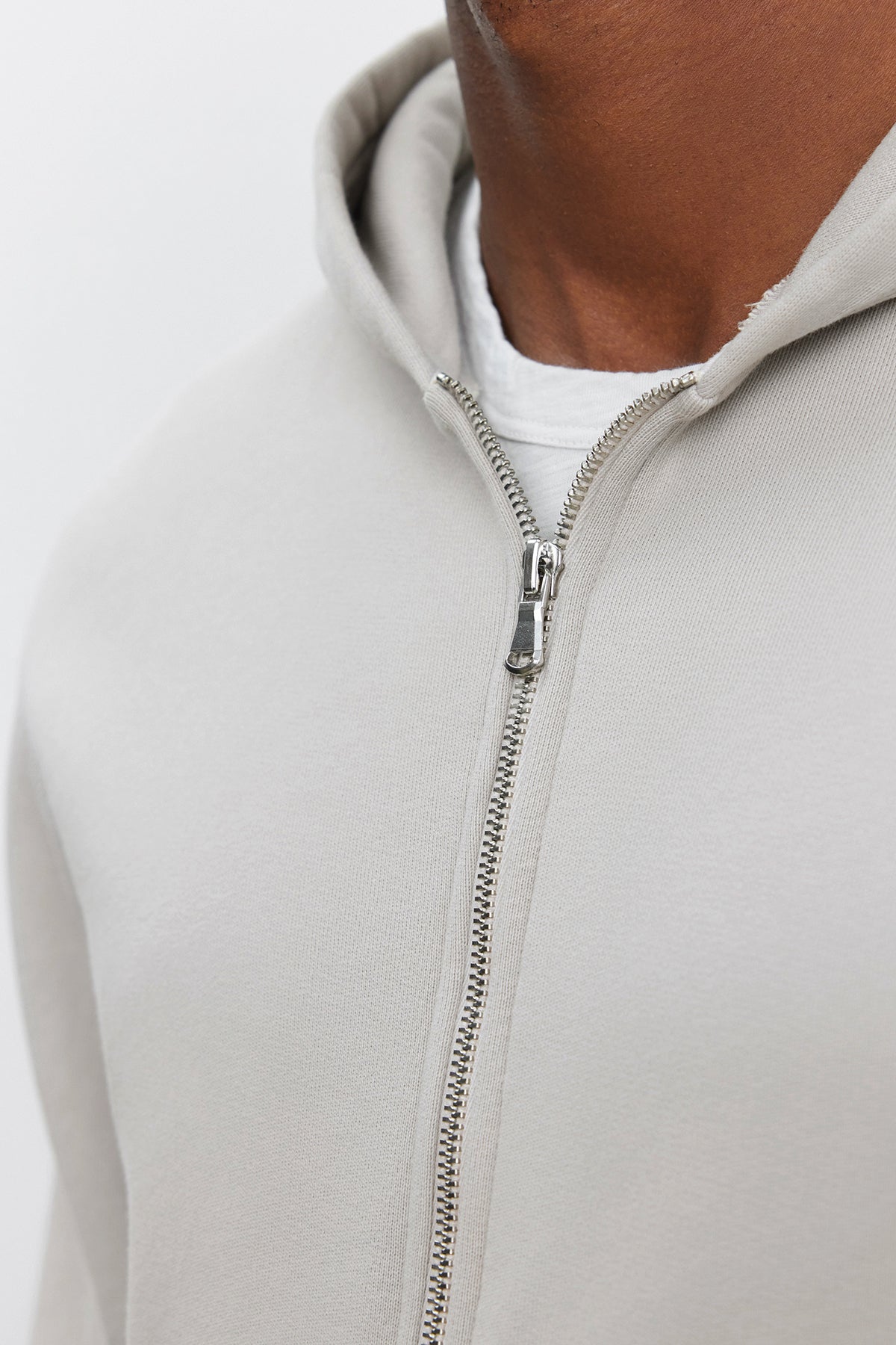   Close-up of a person wearing the REN HOODIE from Velvet by Graham & Spencer, a light gray, cotton fleece, full-zip hoodie over a white t-shirt, focusing on the zipper and chest area. 