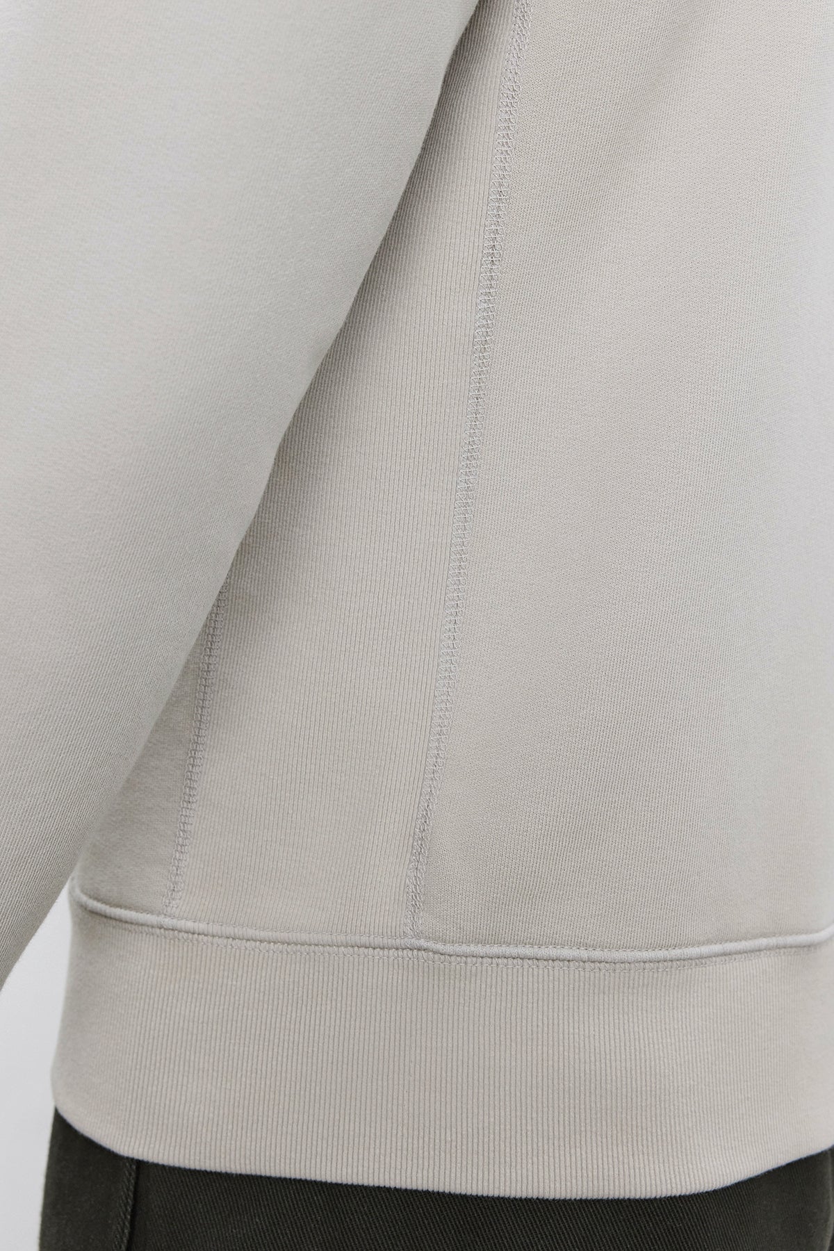 Close-up of the side seam and lower part of a light beige REN HOODIE by Velvet by Graham & Spencer, made from cotton fleece, with the hemline visible, worn over dark green pants.-37893921079489