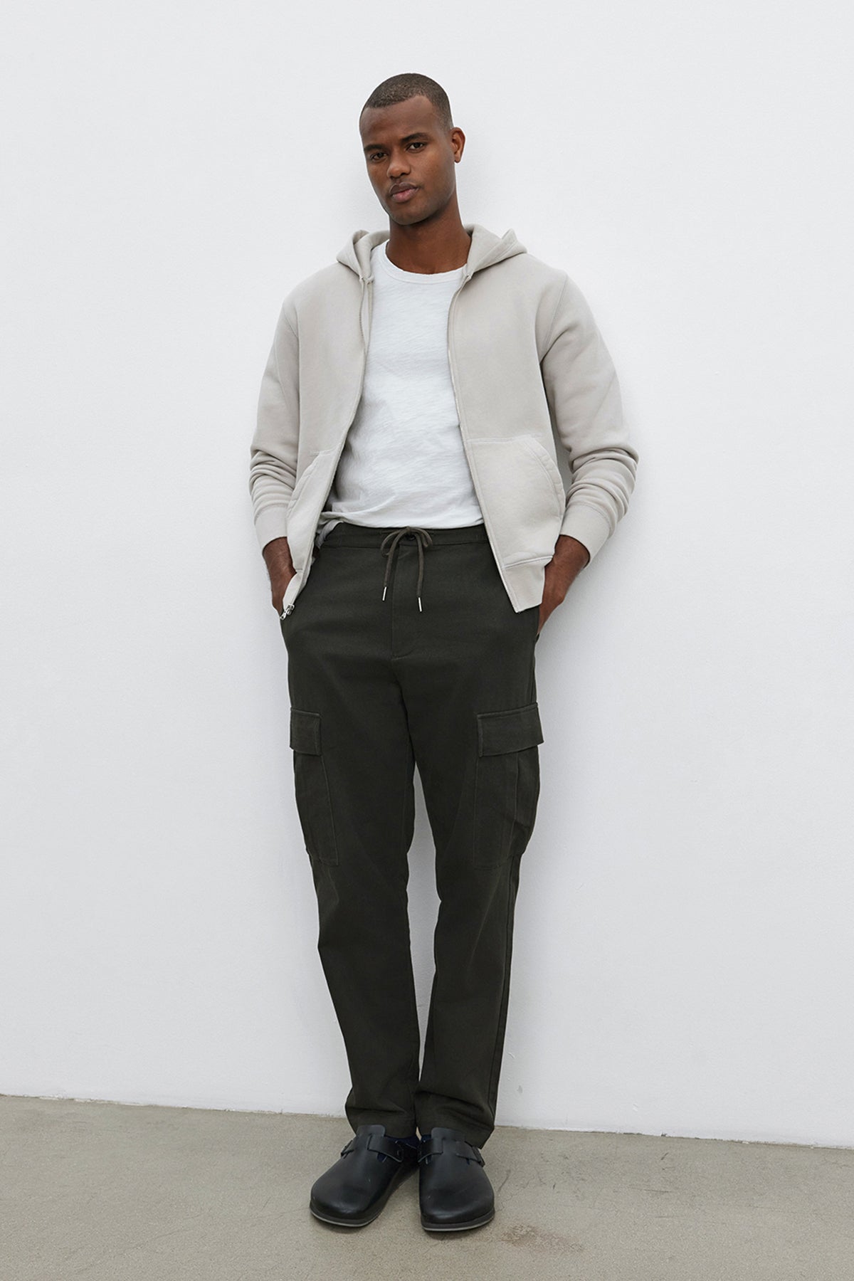 A man stands against a white wall wearing the REN HOODIE by Velvet by Graham & Spencer, a white t-shirt, dark green cargo pants, and black shoes, with his hands in his pockets.-37893921013953