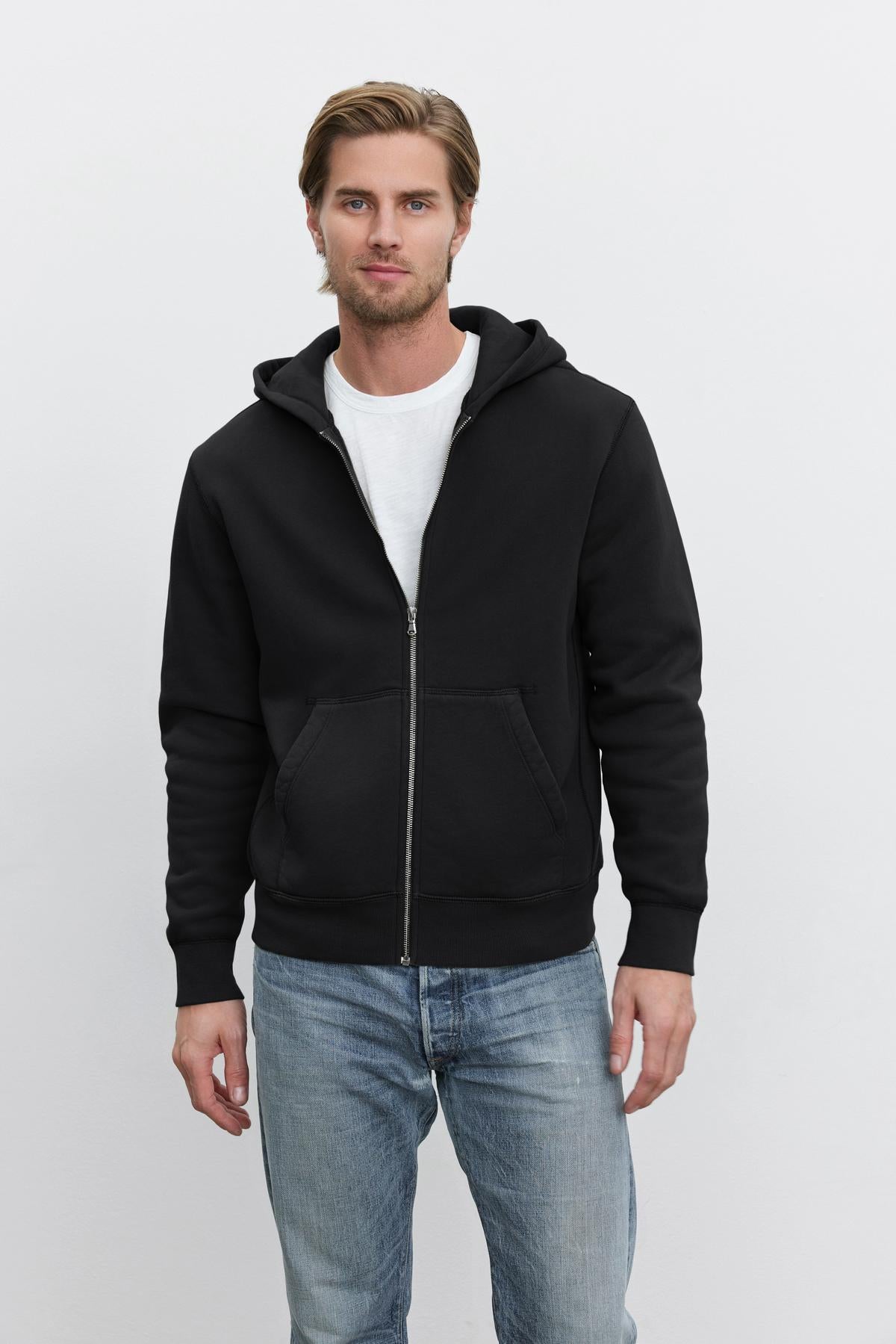   A man wearing a black REN HOODIE by Velvet by Graham & Spencer over a white t-shirt and blue jeans stands against a plain white background, giving off a subtle vintage look. 