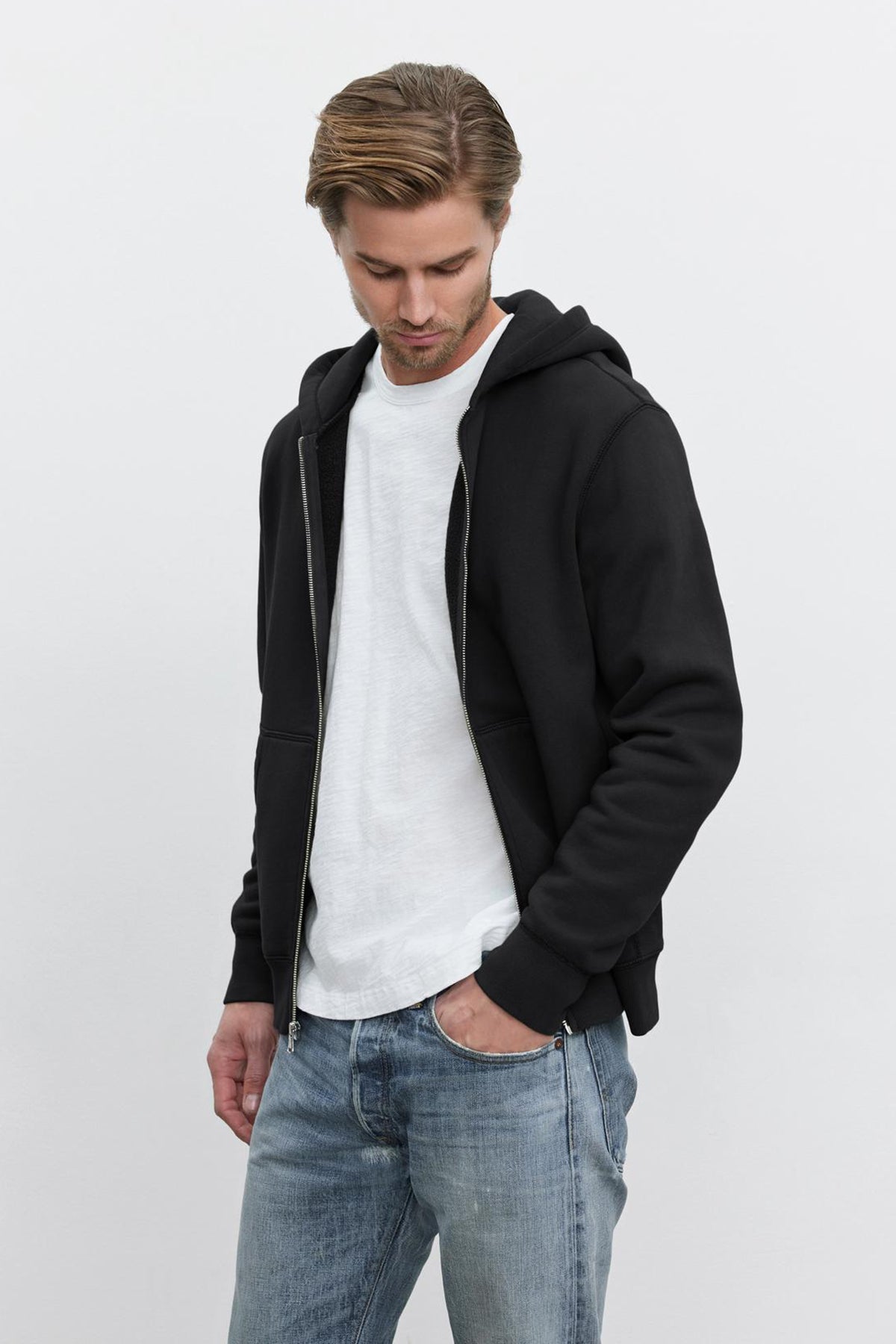   A man wearing the REN HOODIE from Velvet by Graham & Spencer over a white T-shirt and blue jeans looks down with his hands in his pockets, the vintage look enhanced by the soft cotton fleece material. 