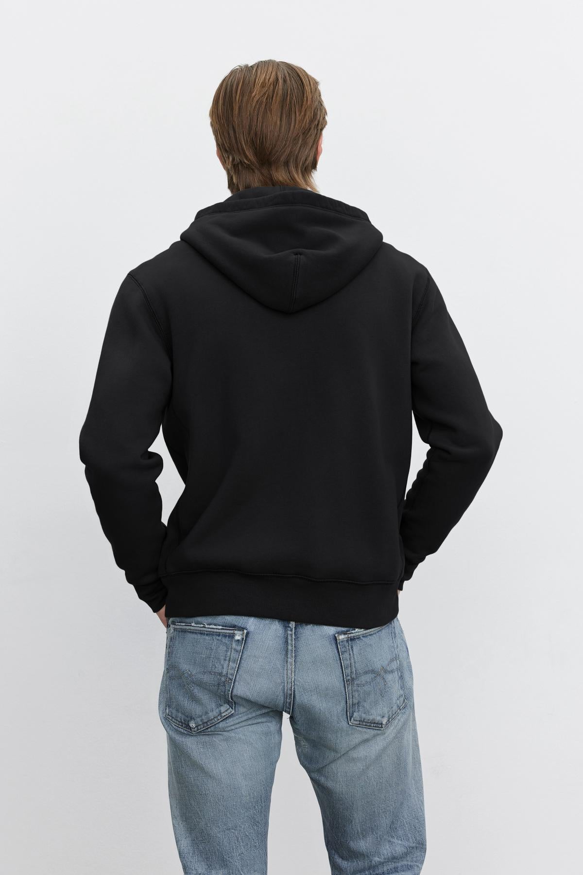   A person wearing the REN HOODIE by Velvet by Graham & Spencer, a vintage-look black full-zip hoodie, and blue jeans stands with their back to the camera against a white background. 