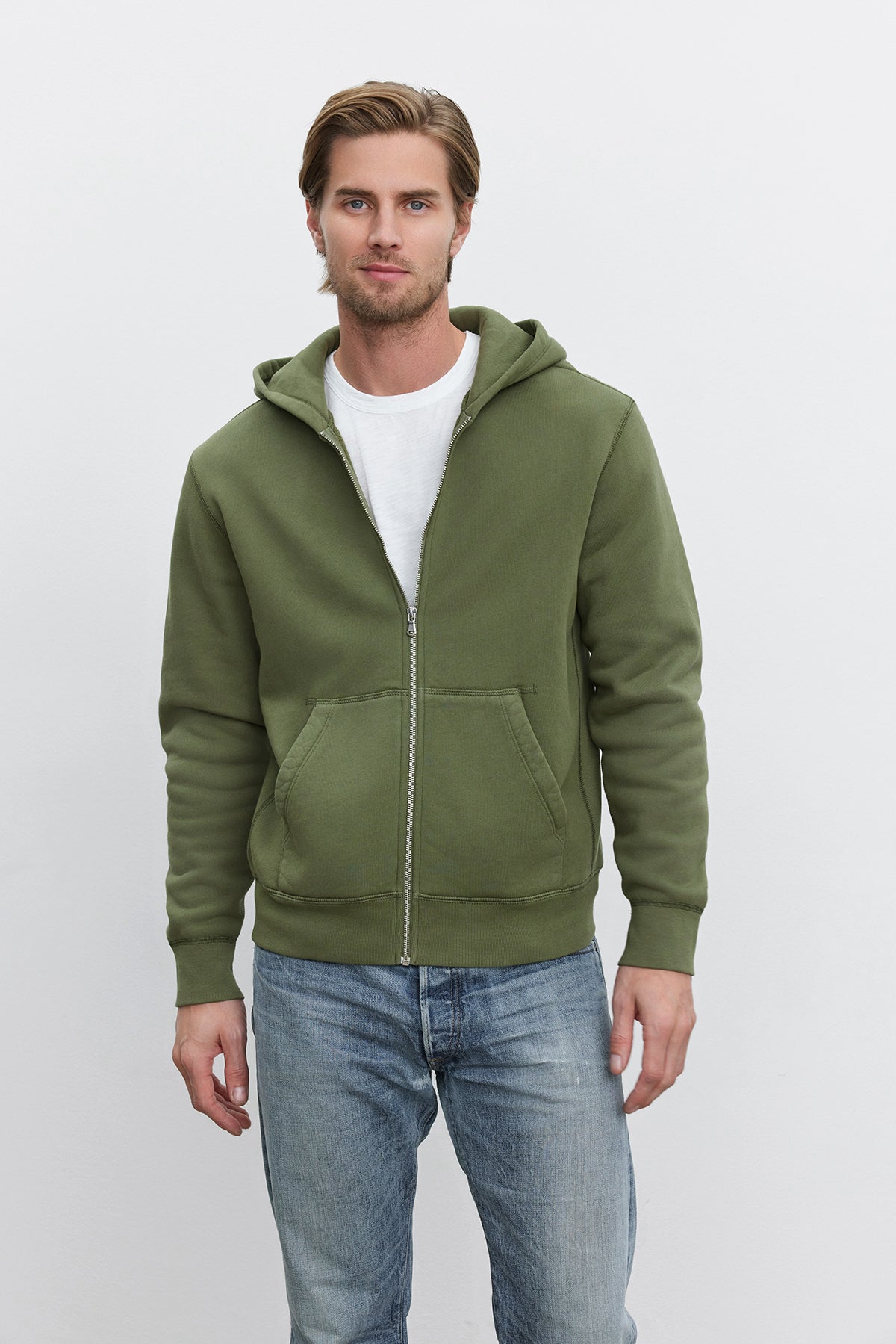 A man with short hair is standing against a plain background, wearing a white T-shirt, blue jeans, and an olive green REN HOODIE with a vintage look from Velvet by Graham & Spencer.-37893745737921