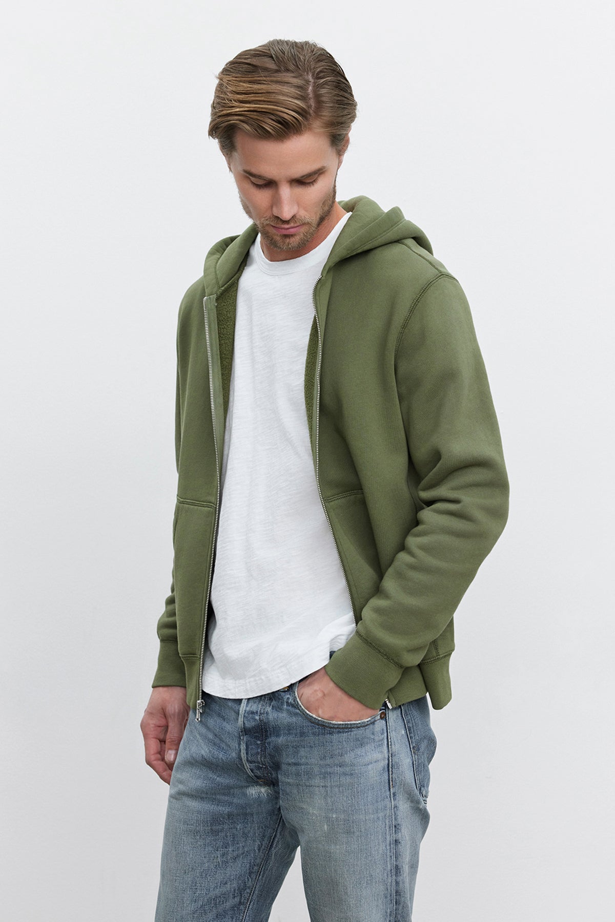 A man wearing the REN HOODIE from Velvet by Graham & Spencer, a green full-zip cotton fleece hoodie, over a white T-shirt and blue jeans stands against a plain background, looking down and exuding a vintage look.-37893745803457