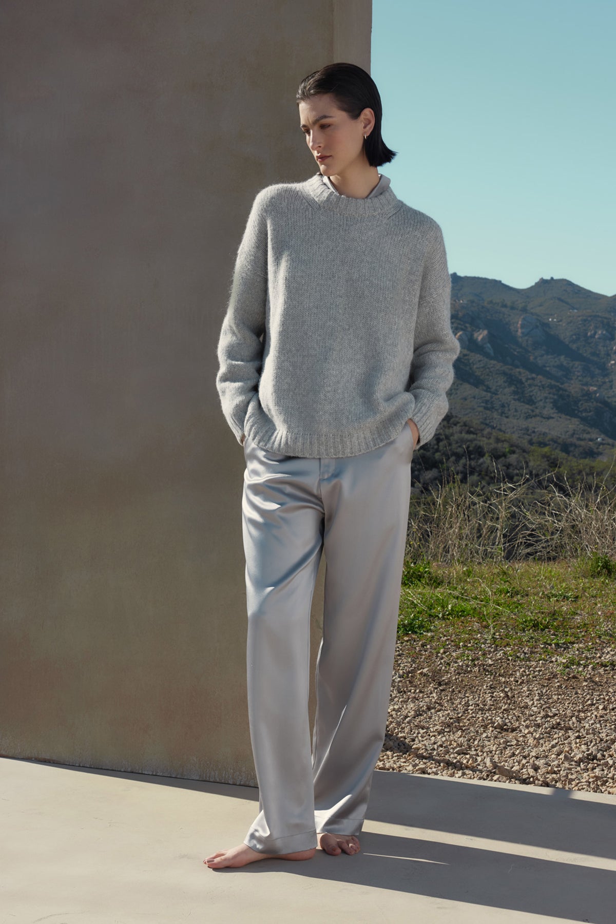   A person with short hair wearing a gray sweater and light-colored MODESTO SILK CHARMEUSE PANTS by Velvet by Jenny Graham stands barefoot outdoors, with their hands in their pockets, against a backdrop of mountains and clear sky. 