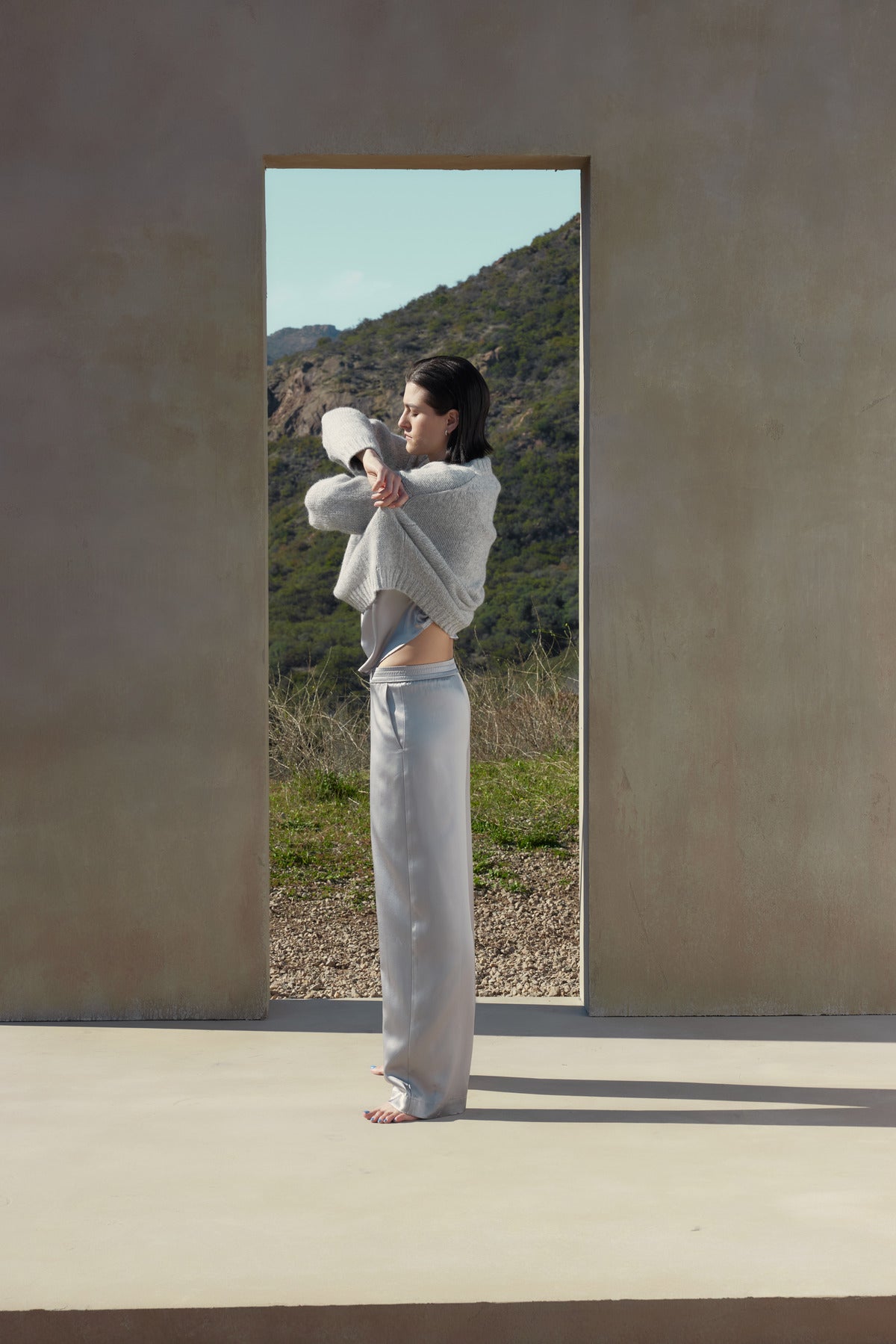   A person stands on a cement platform in front of a rectangular opening in a wall, stretching their arm across their body, clad in MODESTO SILK CHARMEUSE PANT by Velvet by Jenny Graham, with scenic hills in the background. 