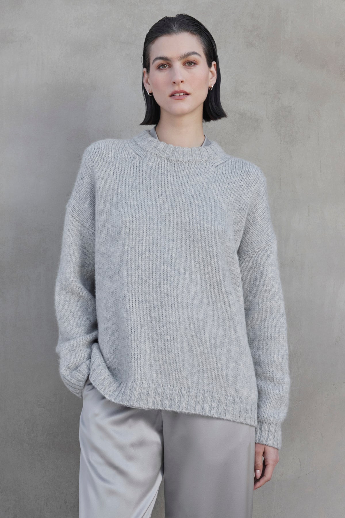 A person with short dark hair wears the CAMBRIA ALPACA SWEATER by Velvet by Jenny Graham, along with light gray pants, standing against a concrete wall.-37659958608065