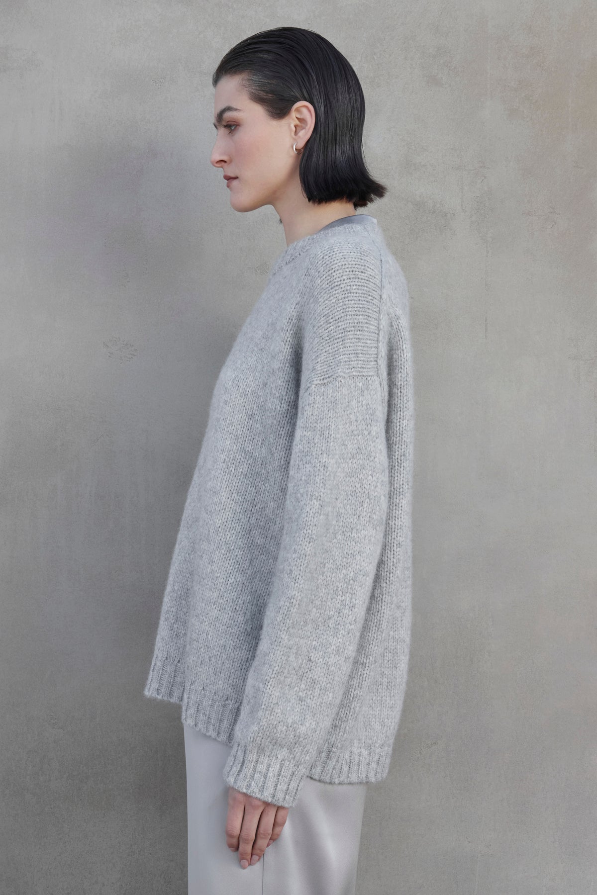   A woman with short dark hair stands against a gray background, wearing the CAMBRIA ALPACA SWEATER by Velvet by Jenny Graham and light pants, facing left in profile view. 